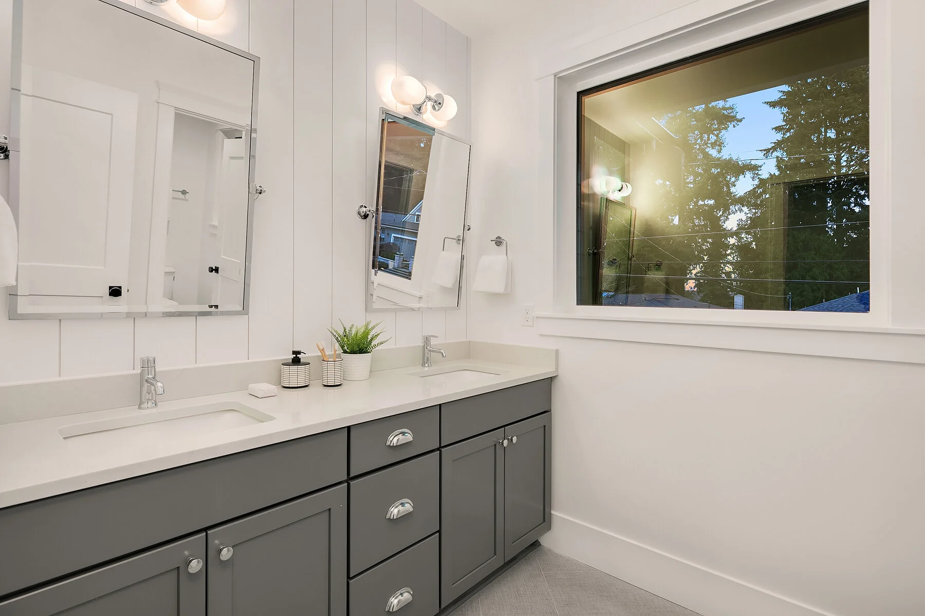 Modern bathroom with dual sinks, grey cabinets, white countertop, large mirror, and window showing trees and evening sky.