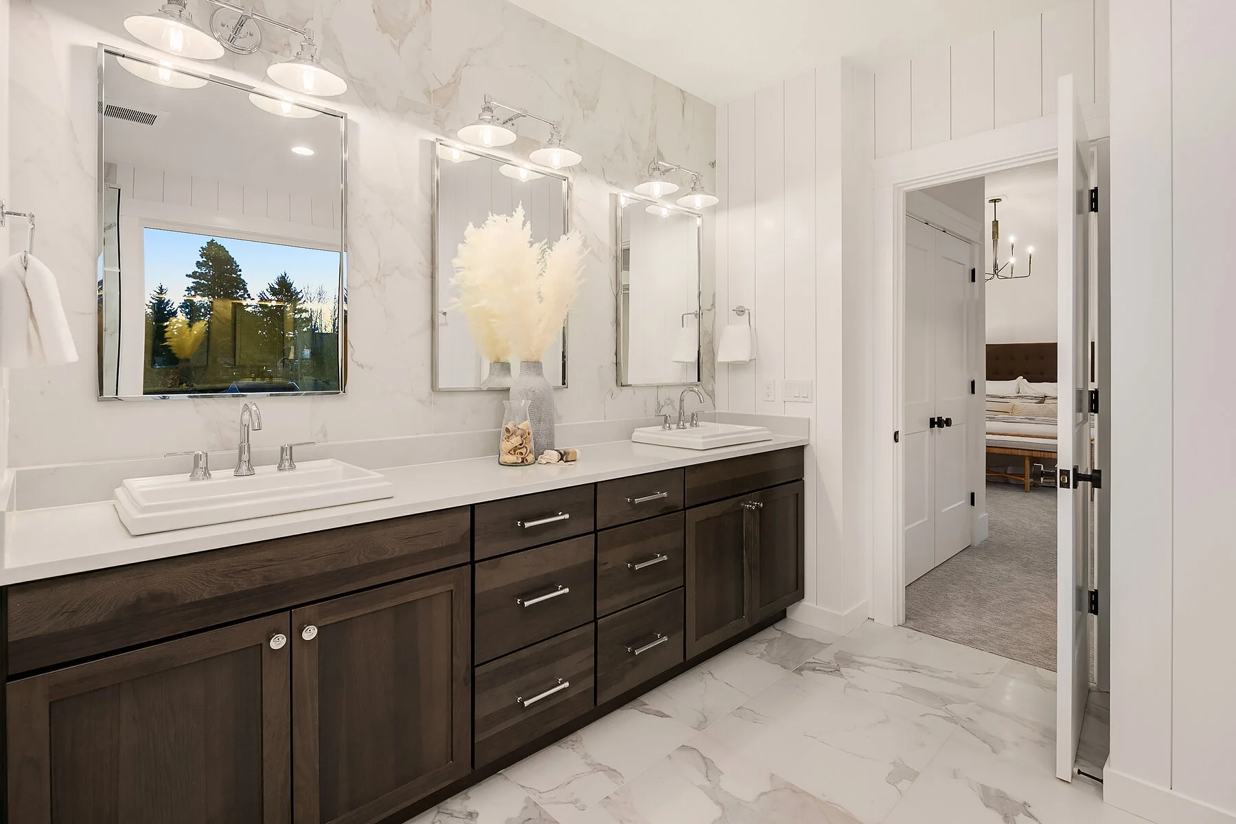 A modern bathroom with dual sinks, large mirrors, and a dark wood vanity. Decor includes a vase with pampas grass and seashells. Next to the vanity is a door leading to a bedroom with a bed and chandelier.
