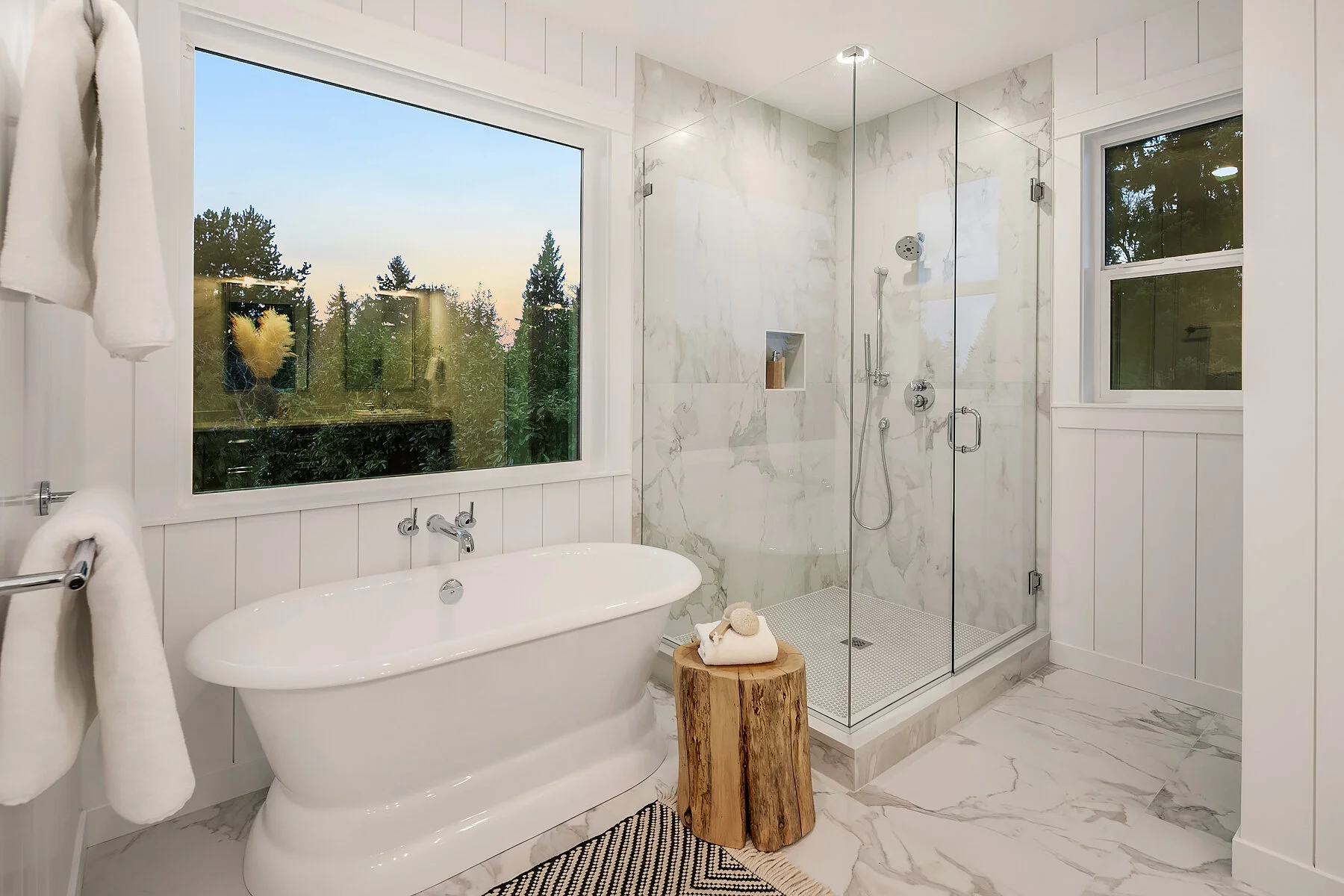 Bright bathroom with white paneling, a wooden stool with towel and bath items, large window showing trees outside, marble floor and shower walls, glass shower enclosure with shower fixtures.