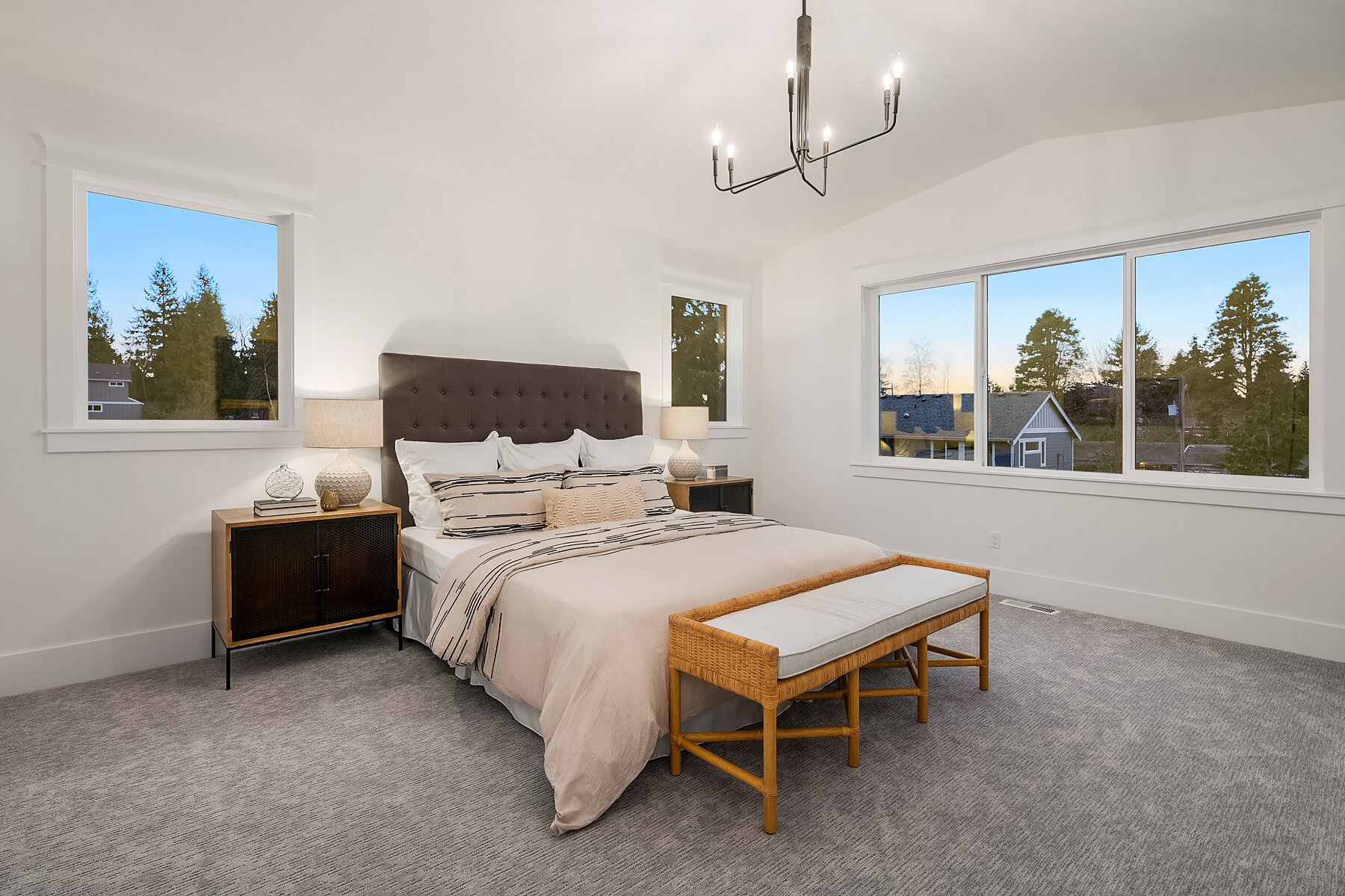A modern bedroom with a bed, two nightstands with lamps, large windows showing trees and houses outside, a bench at the foot of the bed, and a chandelier overhead.