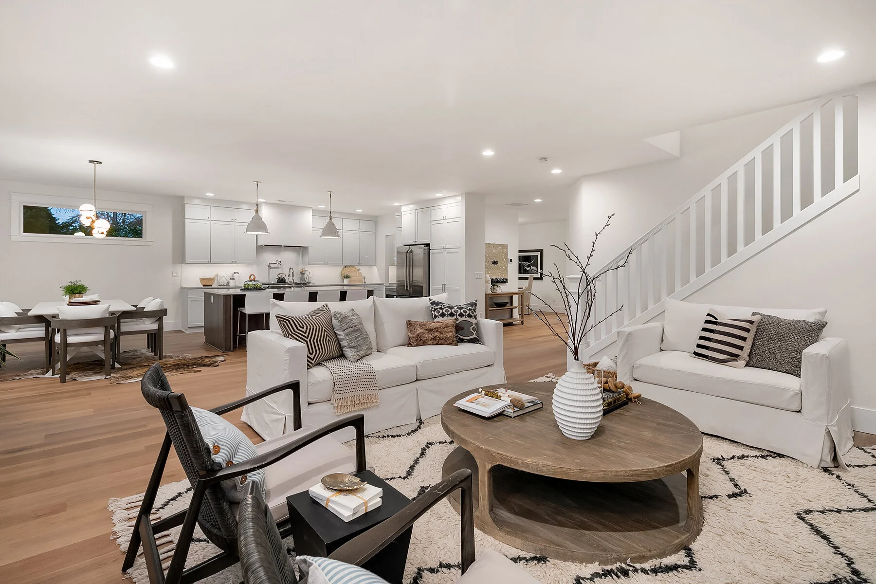 Living room with white sofas, a wooden coffee table, a black armchair, and a white shag rug. The background shows a kitchen with a central island, white cabinets, and stainless steel appliances. There is a stairway to the right and a dining area with