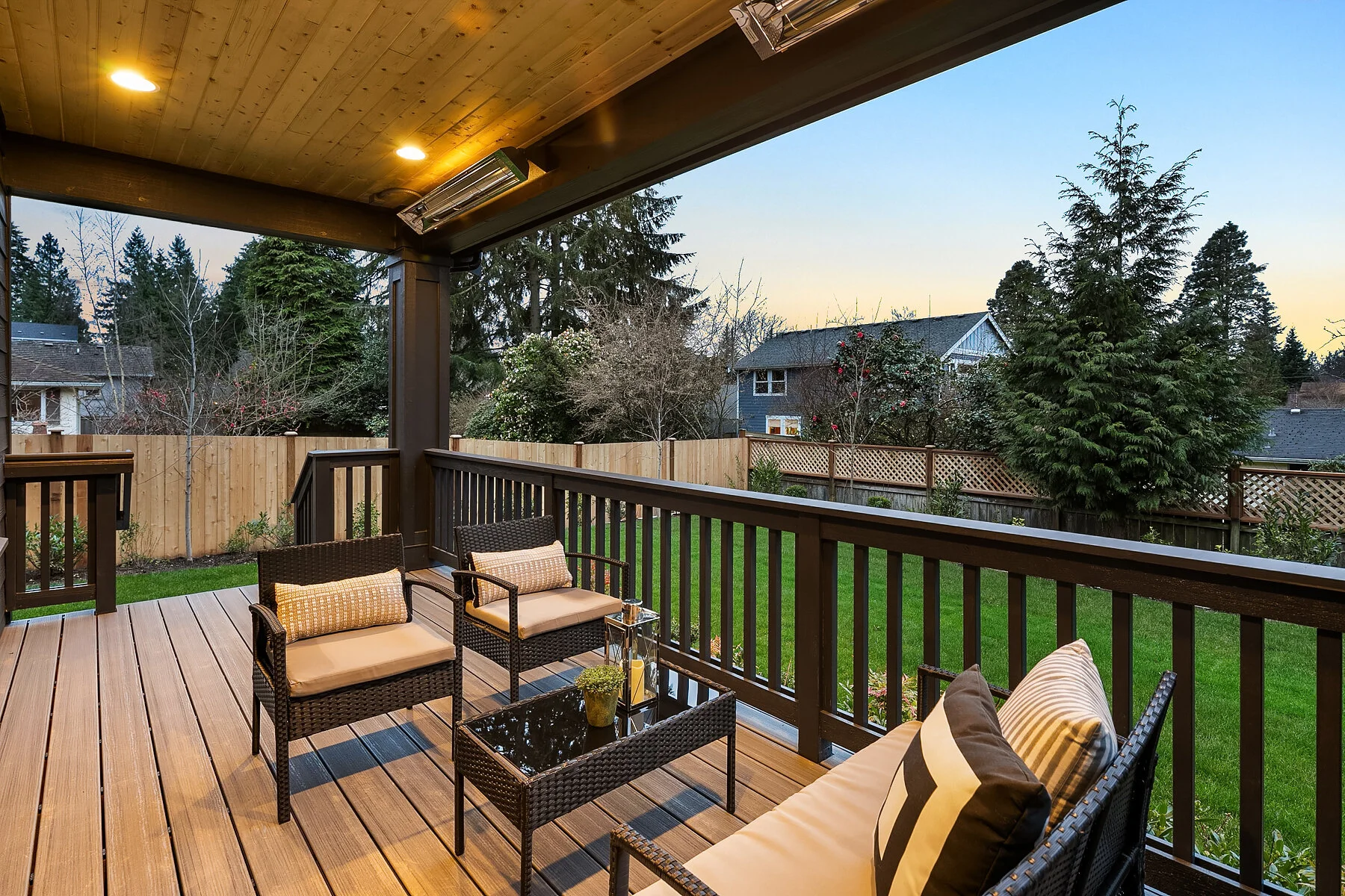A residential backyard with a wooden deck and outdoor furniture, including chairs with cushions and a glass-top table, surrounded by a wooden fence and lush green trees.