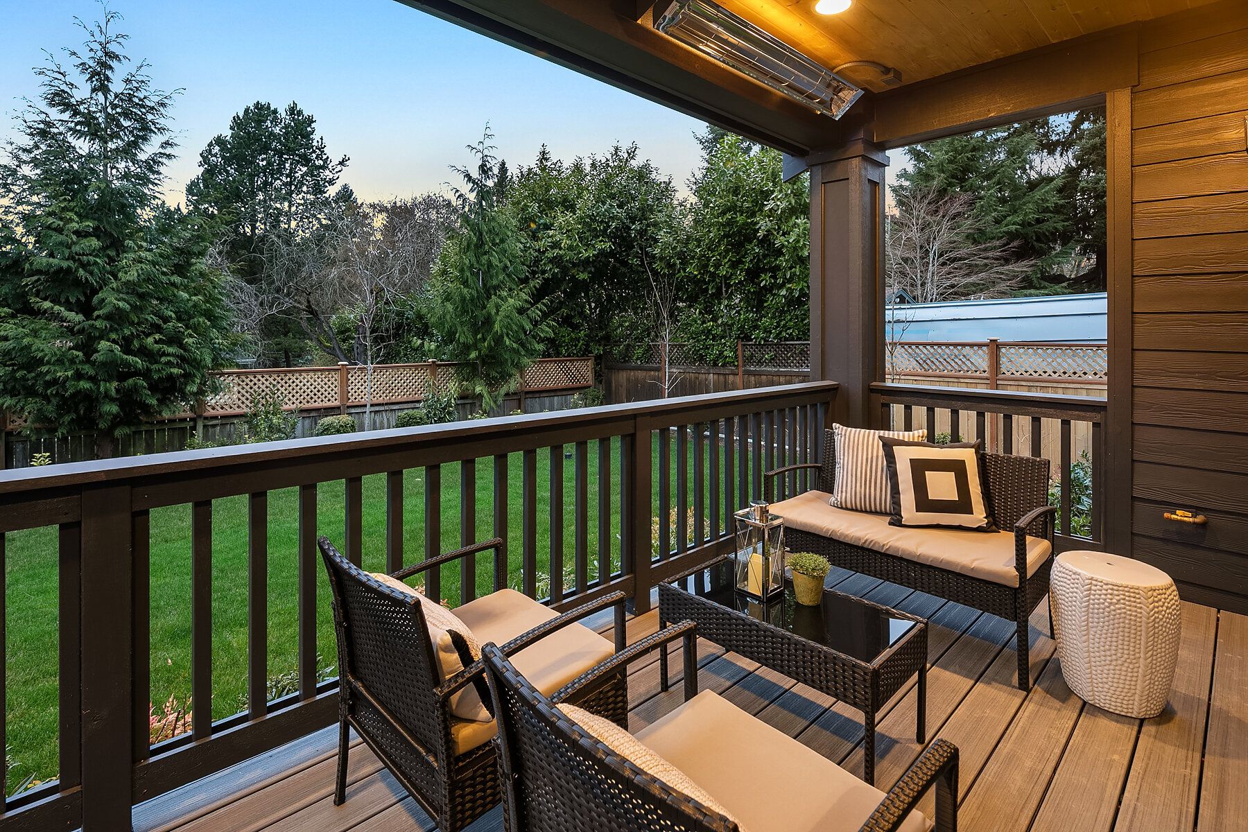 A backyard patio with rattan furniture including a loveseat with pillows, chairs, and a glass-top table, surrounded by trees and a wooden fence.