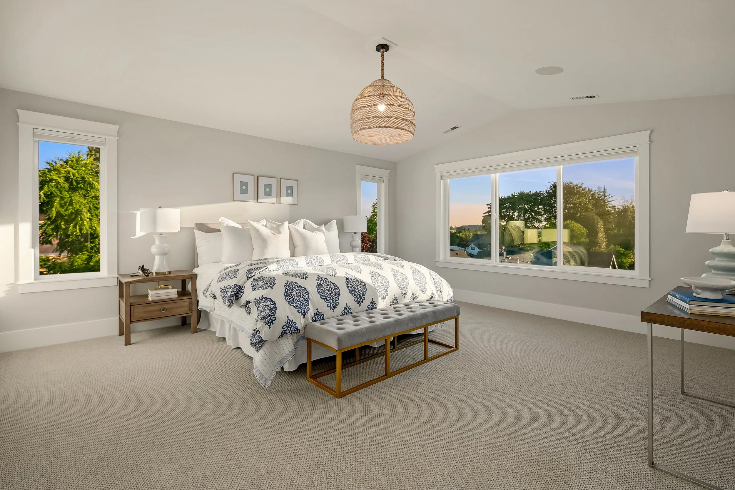 Bright modern bedroom with a large bed, multiple pillows, a wooden nightstand with a white lamp, and large windows showing green trees outside at sunset.
