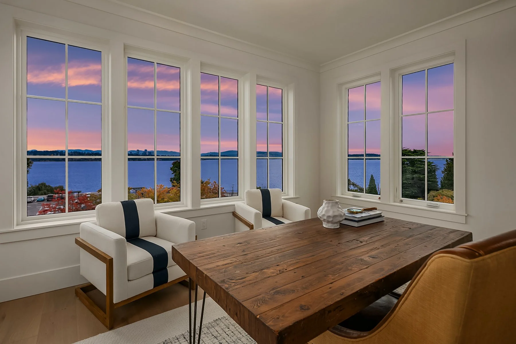 A dining room with three large windows showing a view of a lake and a colorful sunset. The room features a wooden table, two white armchairs with black stripes, and a yellow upholstered chair.