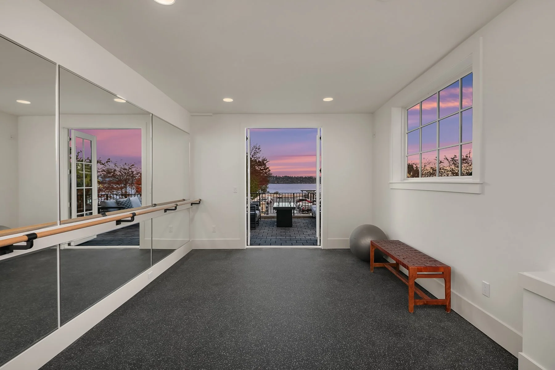 An empty fitness or dance studio with a full-length mirror and ballet barre on the left wall, a window and open door leading to a balcony with outdoor seating and a view of the water and colorful sunset sky, a gray exercise ball, and a wooden bench o