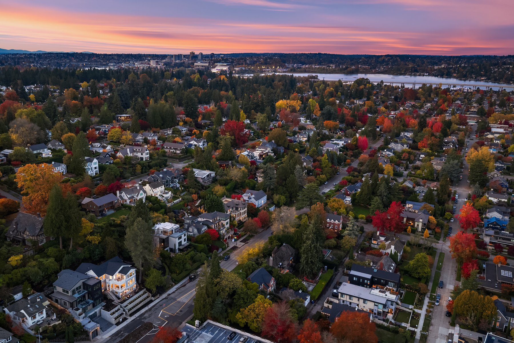 Aerial view of a suburban neighborhood during autumn with colorful fall foliage, houses, and a river in the background at sunset.