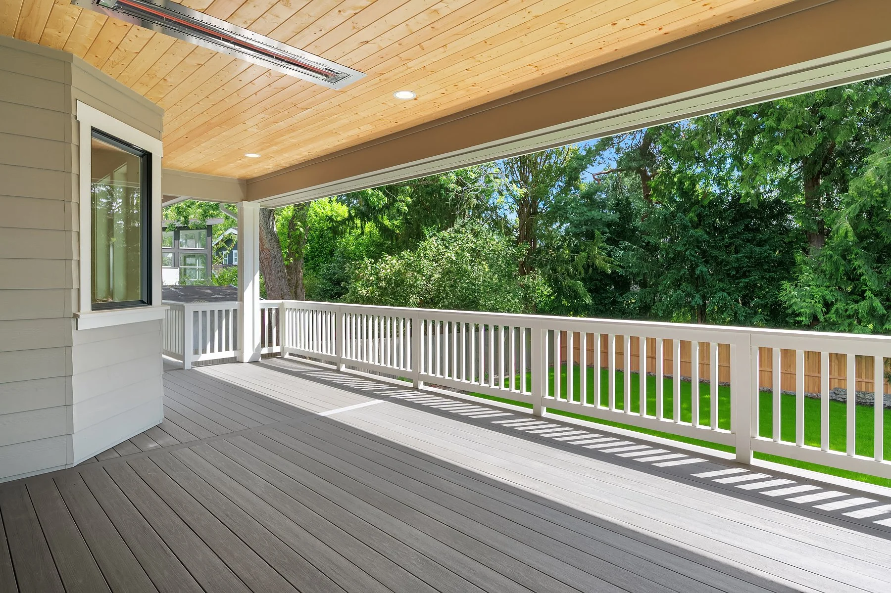 Covered porch with wooden ceiling, white railing, and gray deck, overlooking a backyard with green grass and trees.