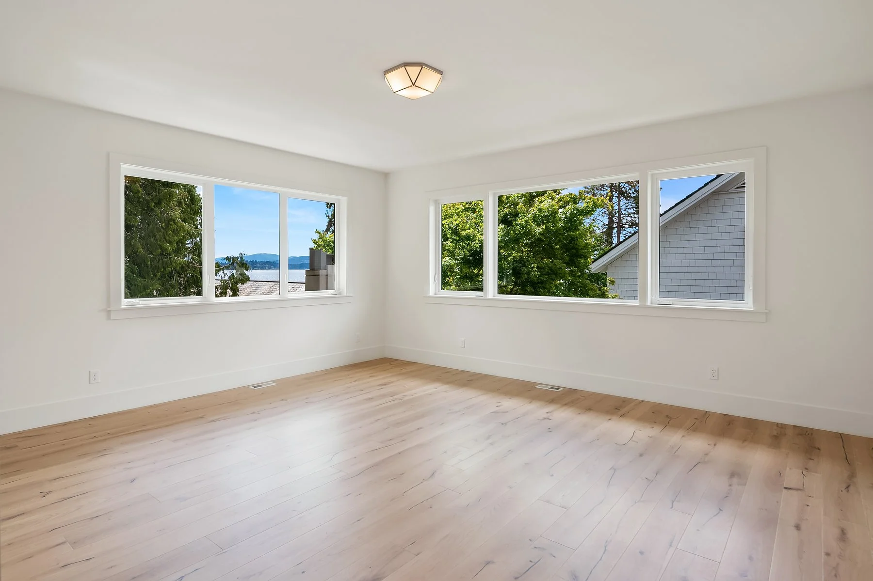 Empty room with white walls, light wood flooring, large windows showing trees, neighboring house, and a view of water and mountains, ceiling light fixture.