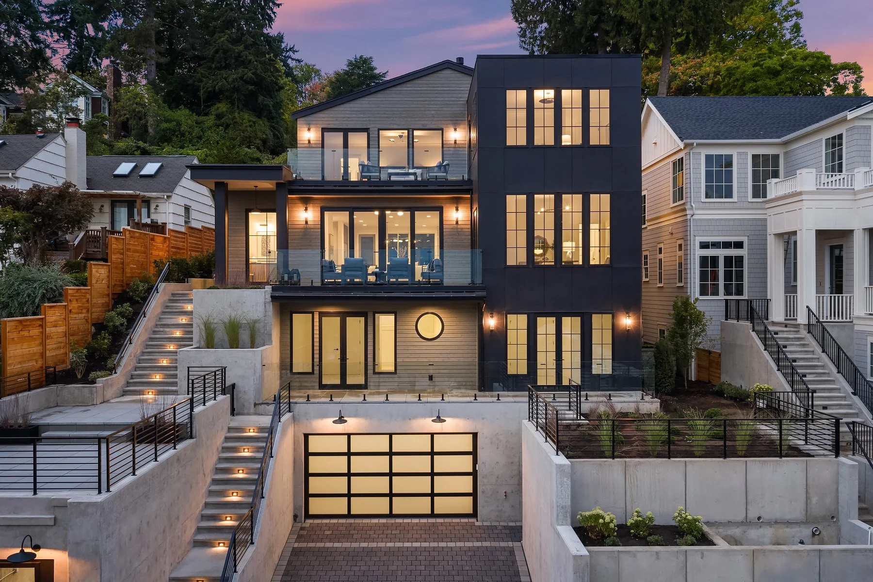 Modern multi-story house with large windows, outdoor terraces, and stairs leading up to the garden, illuminated at dusk.