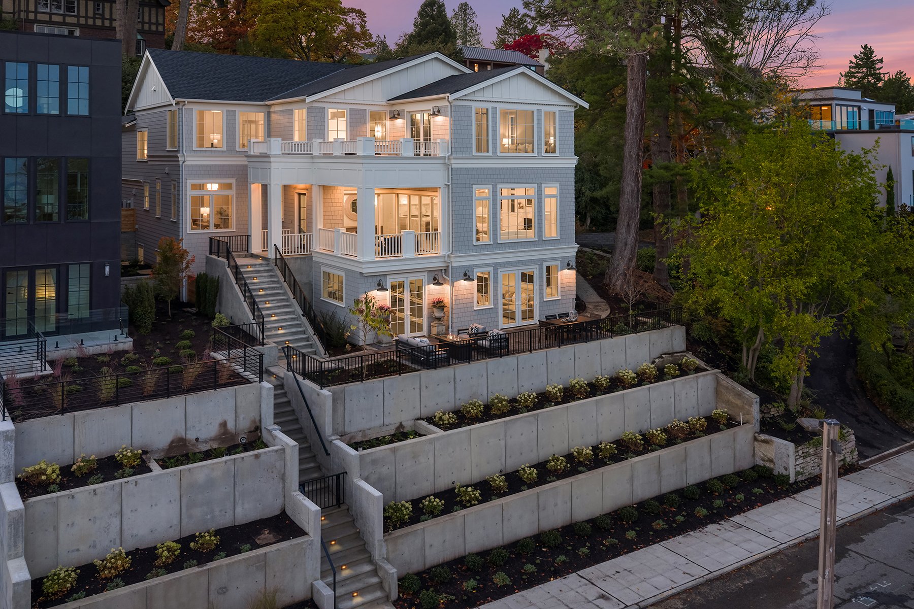 Multi-story house with large windows illuminated from the inside, landscaped garden, and tiered concrete retaining walls with stairs.