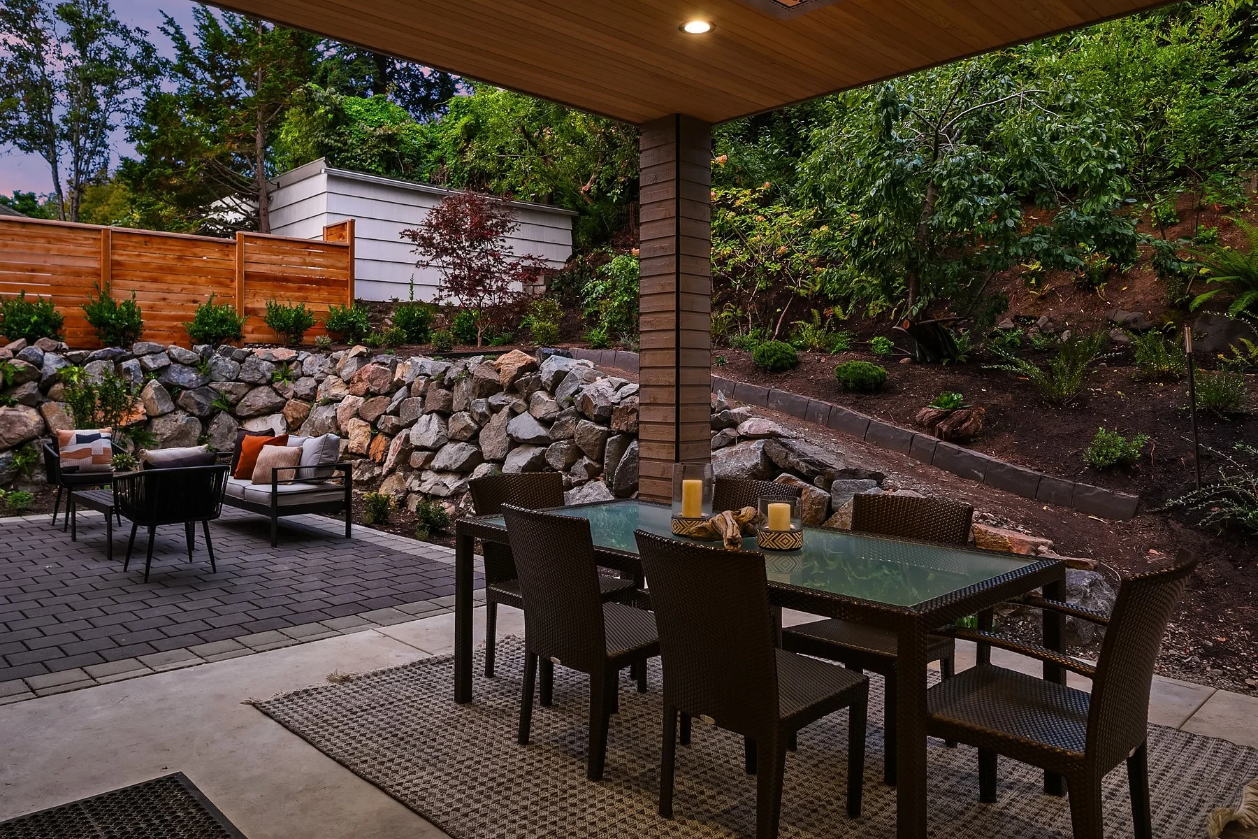 Outdoor patio with a dining table and chairs under a covered area, overlooking a landscaped garden with rocks, plants, and trees.