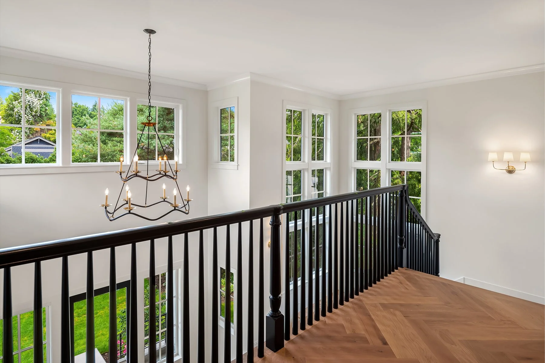 Bright, airy upstairs landing area with large windows showing green trees outside, a chandelier hanging from the ceiling, a wall-mounted sconce light, black wooden staircase railing, and hardwood flooring.