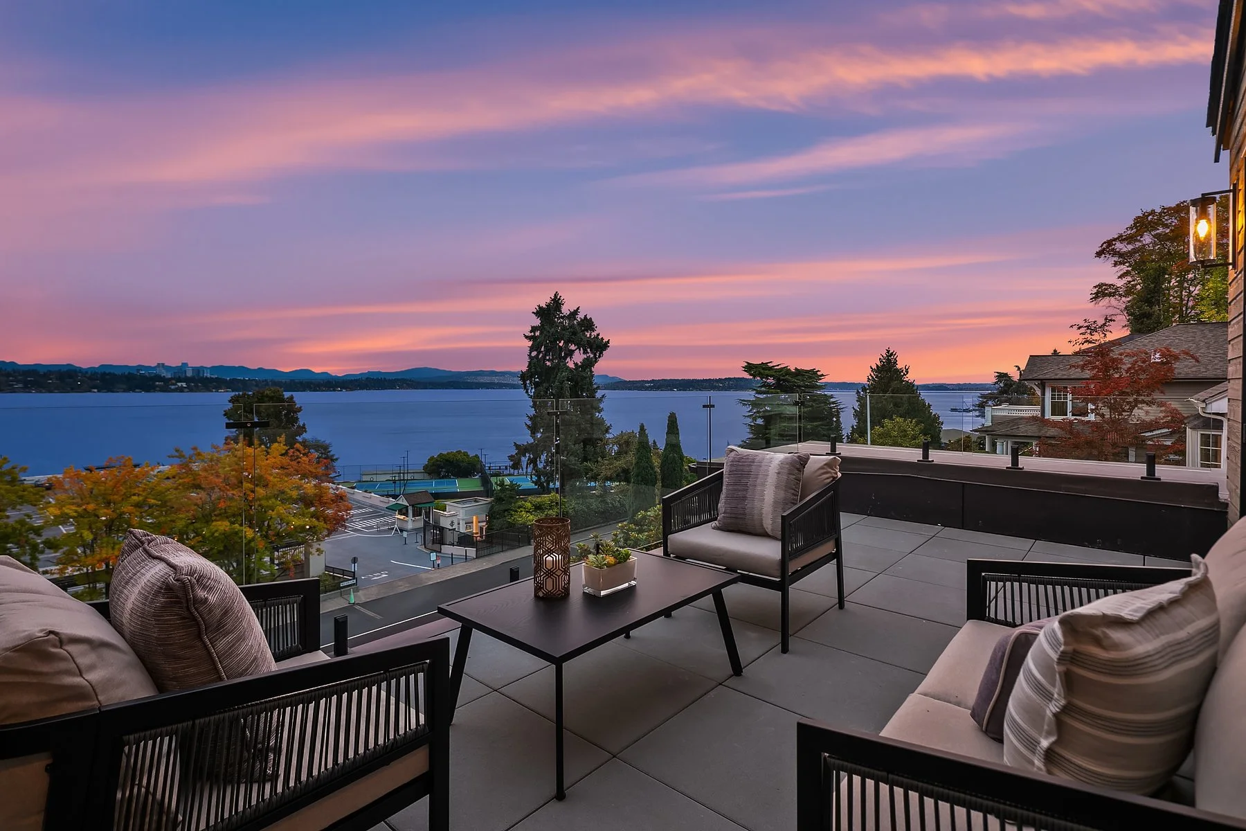A balcony overlooking a body of water at sunset, with outdoor furniture including cushioned chairs and a table decorated with a candle and a small plant.
