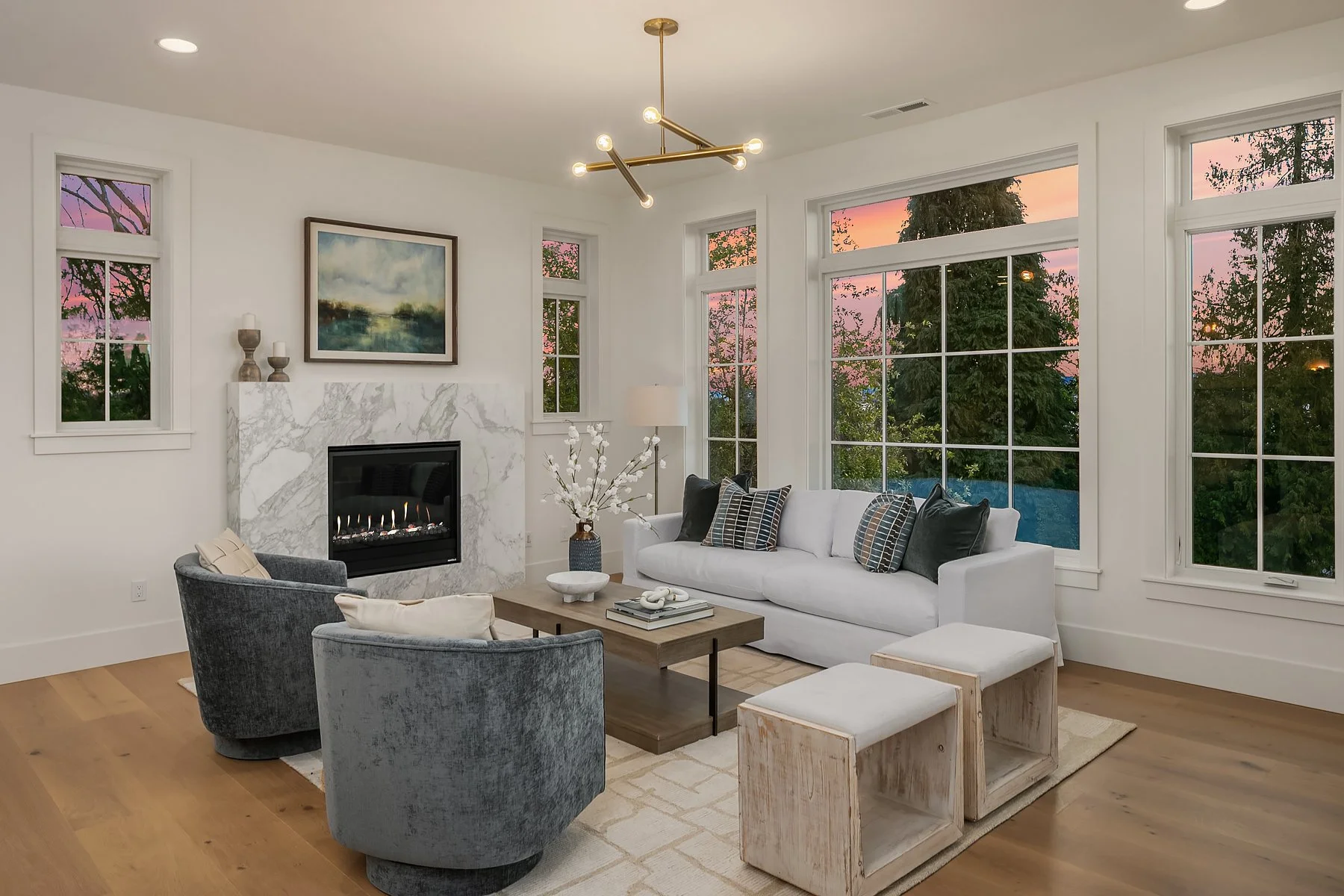 Living room with white walls, large windows showing a sunset, a white sofa with multiple pillows, two gray armchairs, a wooden coffee table, a marble fireplace, and modern light fixtures.