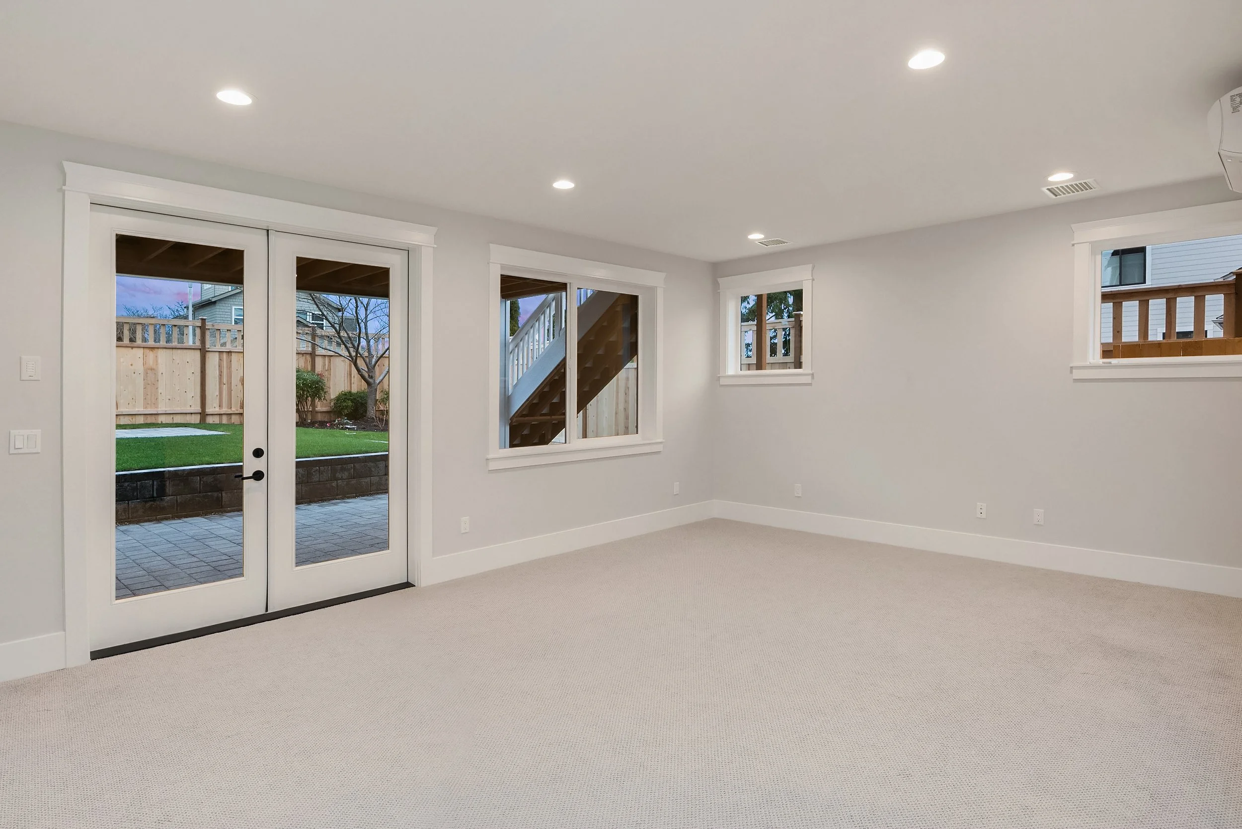 Empty living room with beige carpet, white walls, multiple ceiling lights, and windows showing a backyard with a wooden deck, fence, and trees.