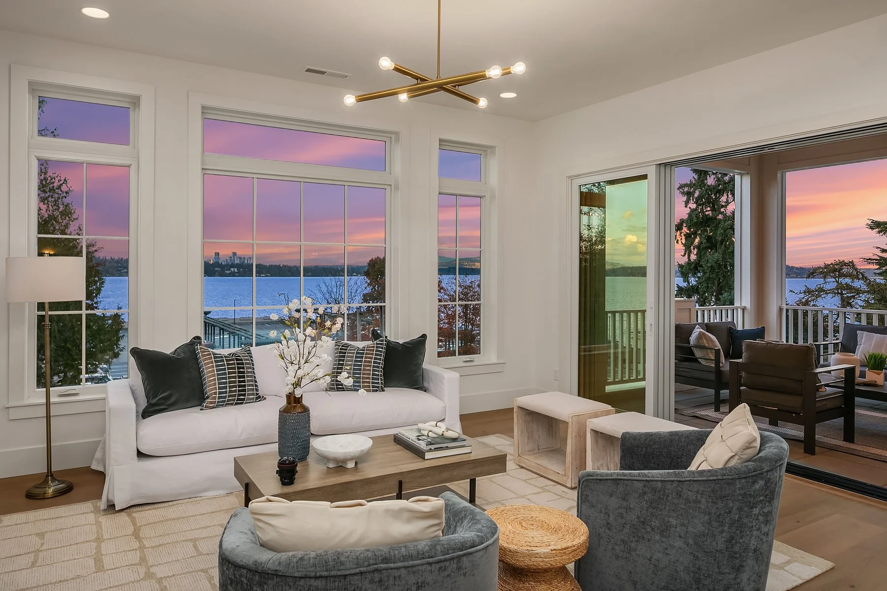 Living room with large windows overlooking a lake and colorful sunset sky, furnished with a white sofa, gray armchairs, wooden coffee table, and patio area.