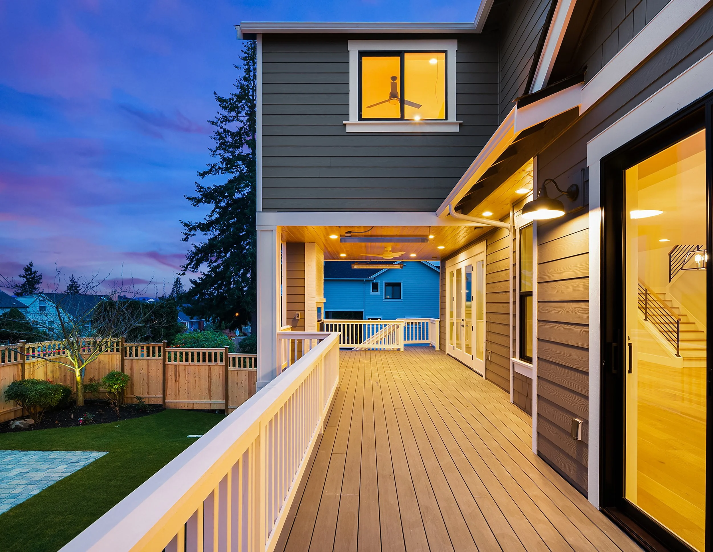A spacious outdoor balcony with wooden flooring, white railings, and a view of a backyard with a tree and a fence, illuminated by outdoor lighting at dusk, attached to a modern house with large glass doors and windows showing a staircase inside.