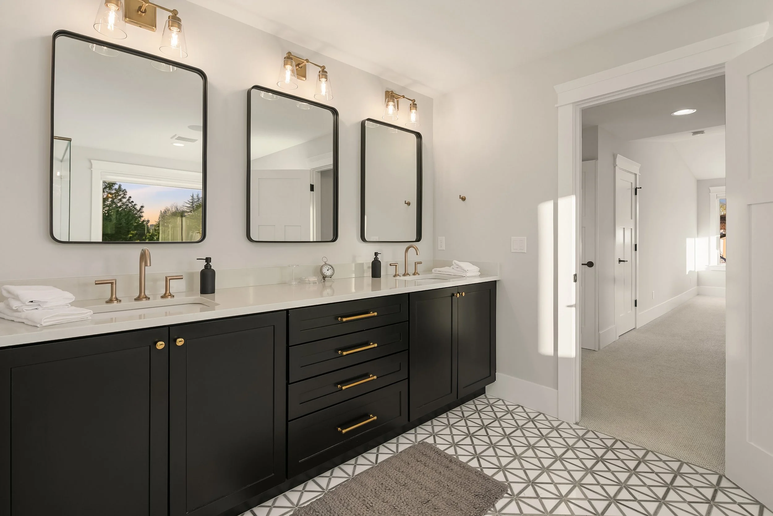A modern bathroom with black vanity, white countertop, three mirrors, and gold fixtures. There are wall-mounted lights above the mirrors and a view of the outdoors through a window reflected in one of the mirrors.