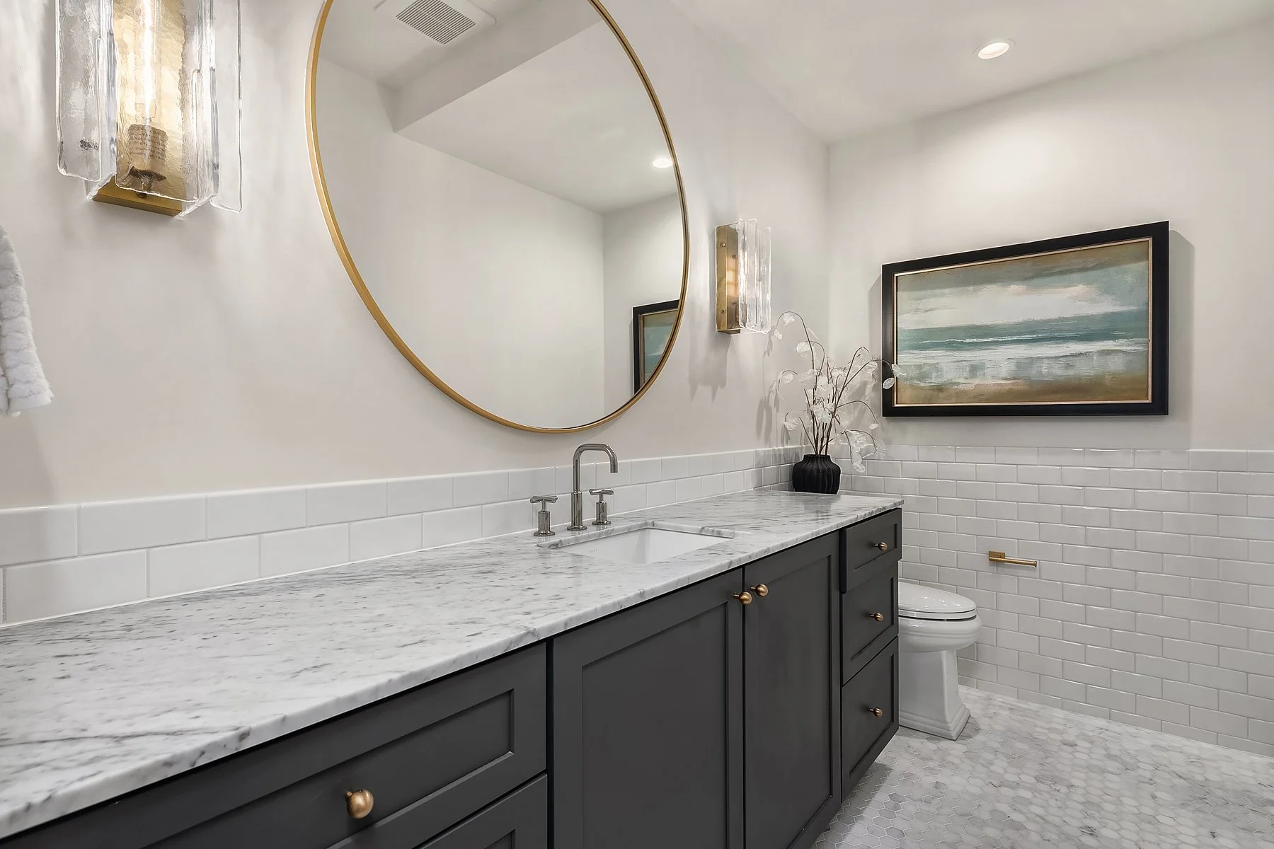 Bathroom with dark gray vanity, white marble countertop, round mirror, wall sconces, painting of ocean, vase with decorative branches, white subway tile walls, toilet, and hexagonal tile floor.