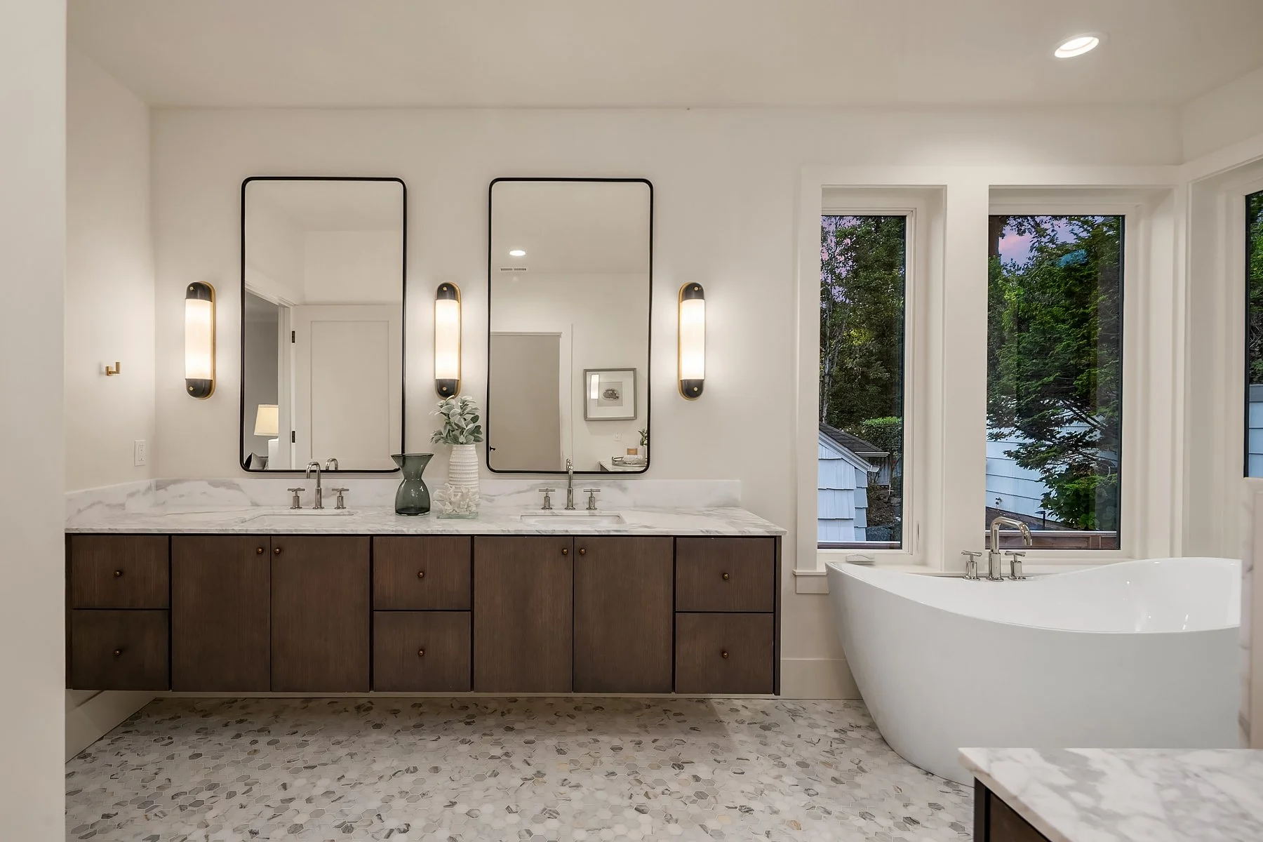 Modern bathroom with double vanity, two large mirrors, wall-mounted lights, a white freestanding bathtub, and large windows showing trees outside.