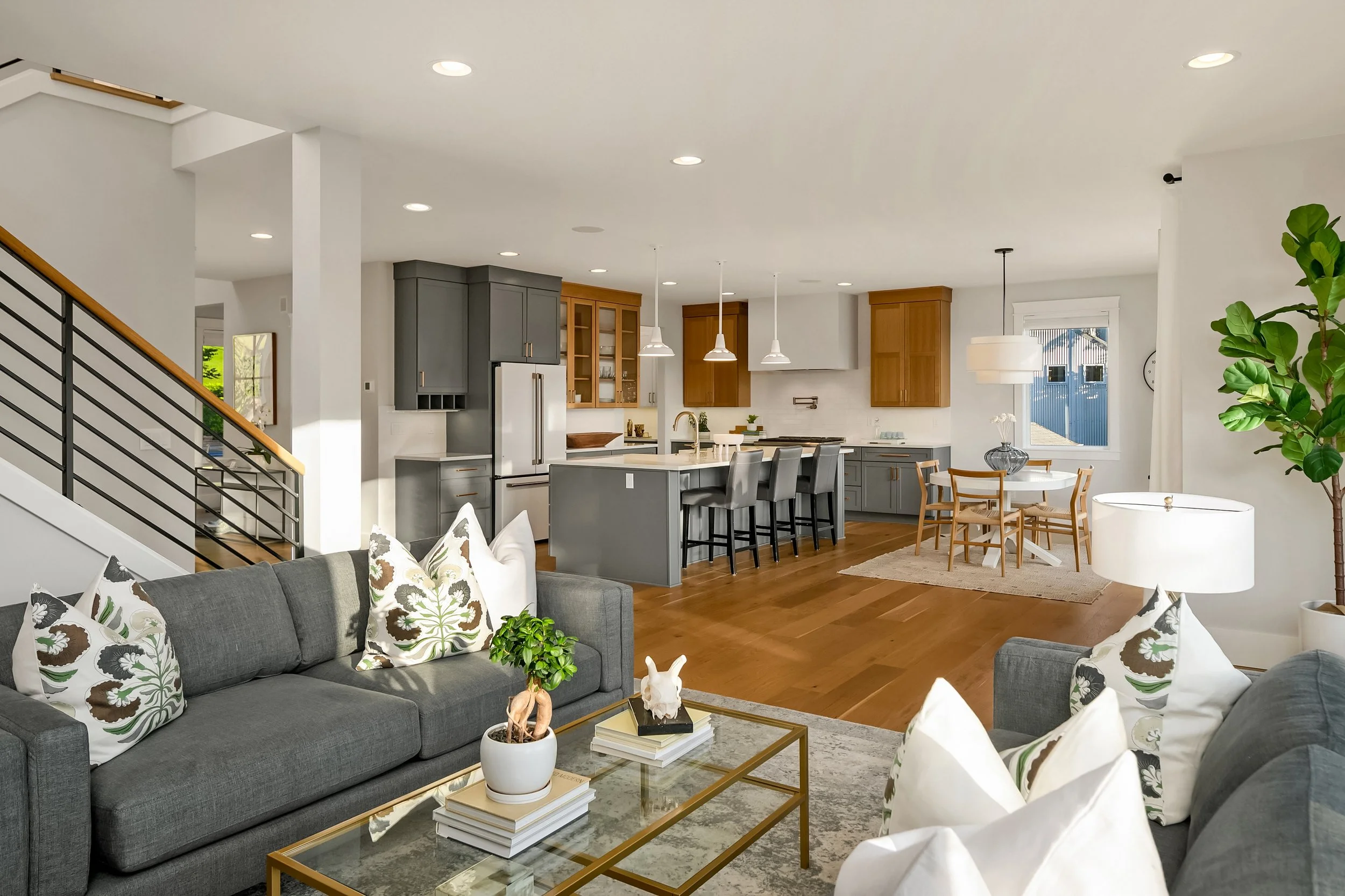Living room with grey sofas, decorative pillows, a glass coffee table, and houseplants, open to a kitchen and dining area with wooden floors and modern decor.