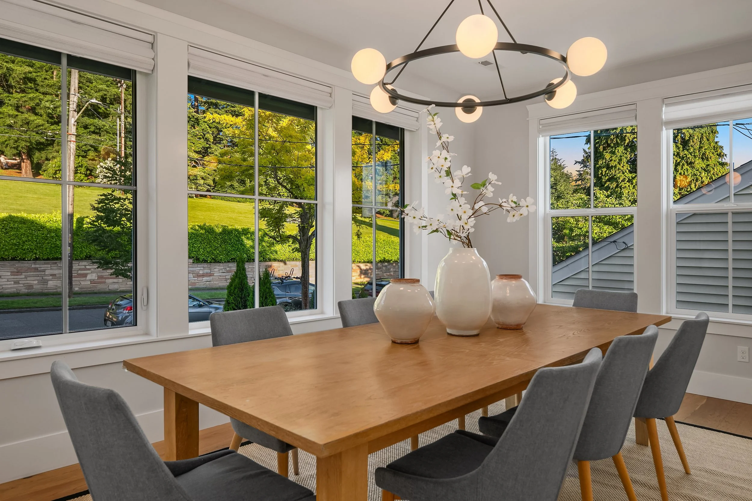 A dining room with large windows showing a green outdoor view, a wooden table with six gray chairs, and decorative vases with flowers, illuminated by a modern circular ceiling light.
