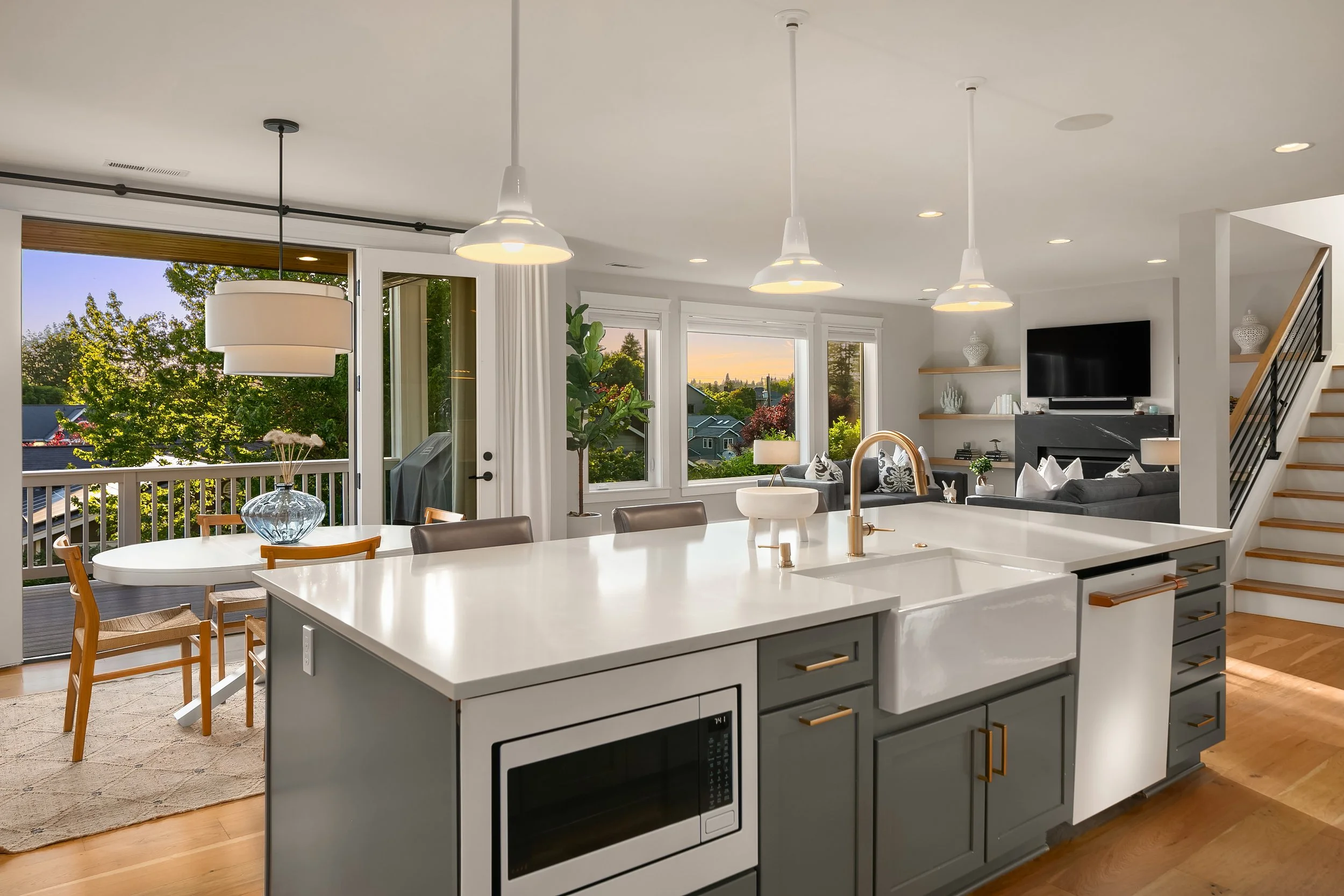 Modern open-concept kitchen and living room with large windows, greenery outside, and a view of the sunset.