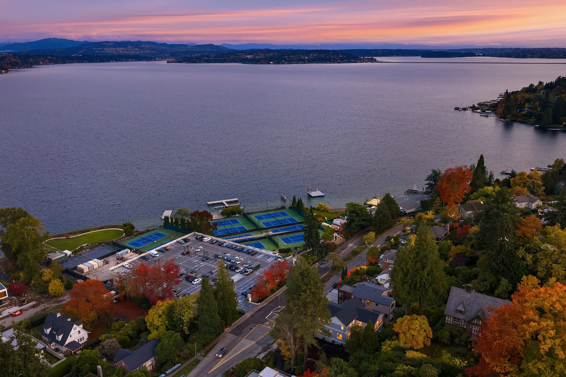 Aerial view of a lakeside neighborhood with houses, colorful trees, and a parking lot with tennis courts near the water, during sunset.