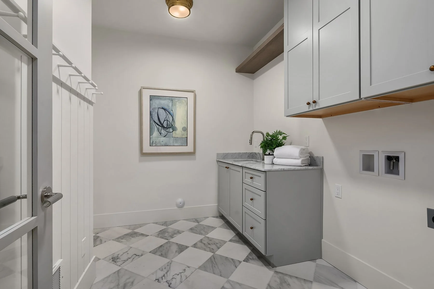 Small modern laundry room with gray cabinets, marble countertop, a sink, a green plant, and folded white towels. The floor has a white and gray checkered tile pattern. There is a framed abstract artwork on the wall.