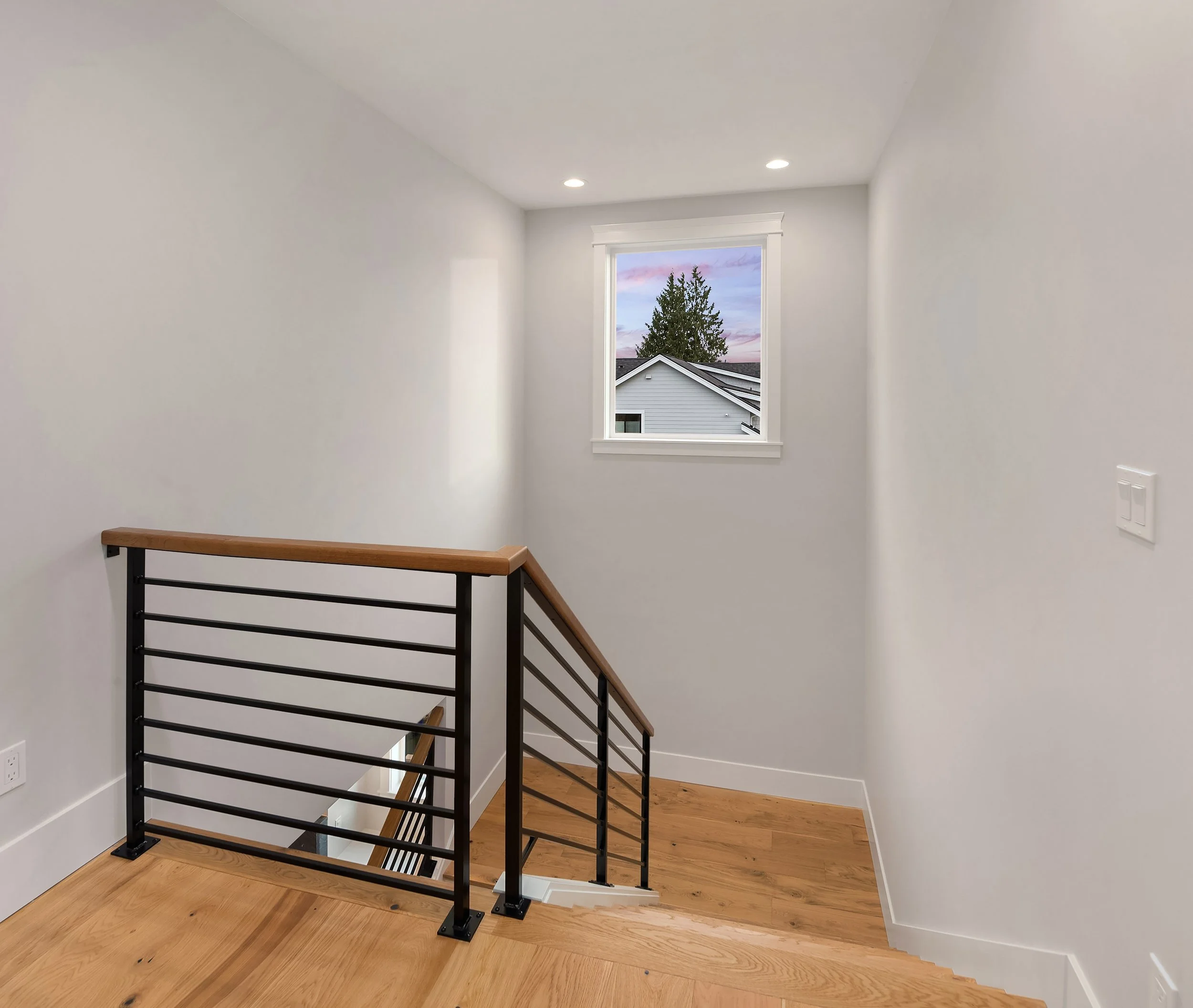 Empty white room with a window showing an outdoor scene, a staircase with black metal railing and wooden handrail, and wooden flooring.