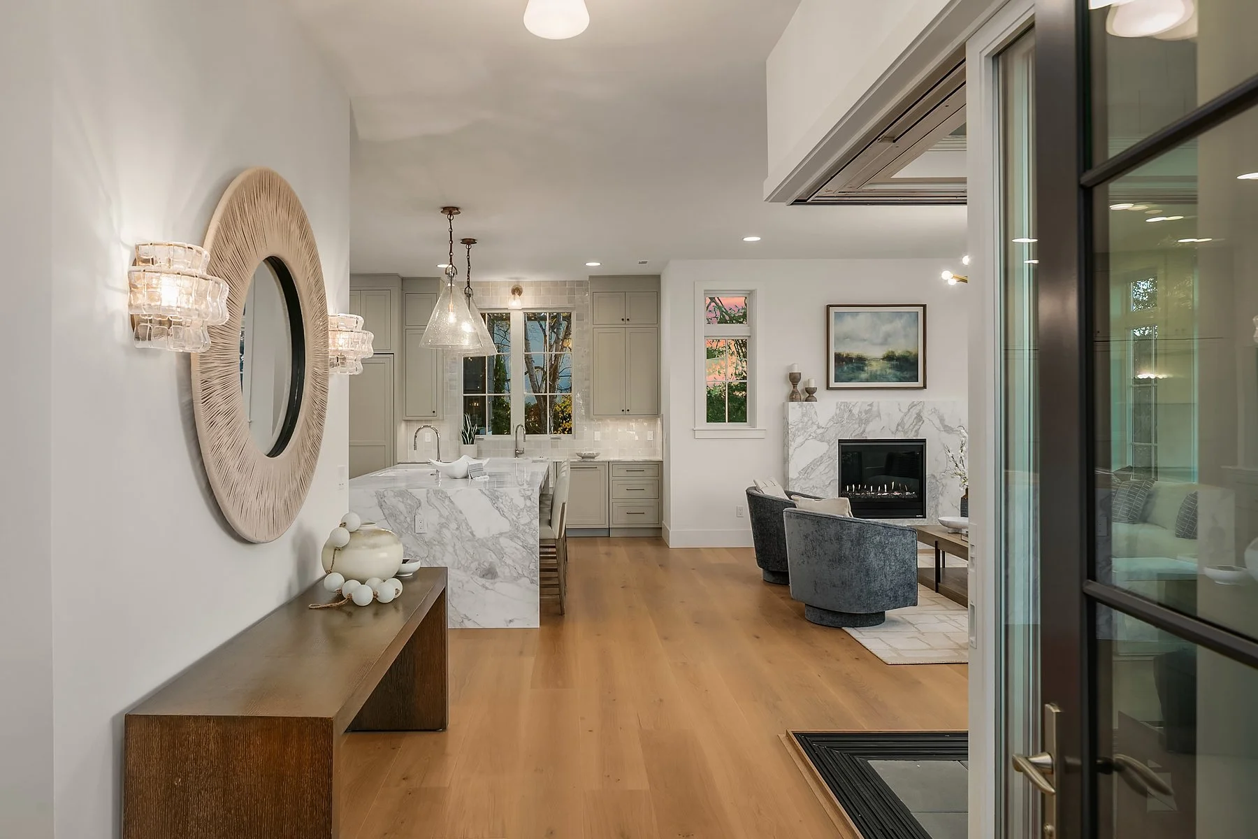 Modern living room and kitchen with white walls, hardwood floors, marble countertops, gray chairs, fireplace, and artwork.