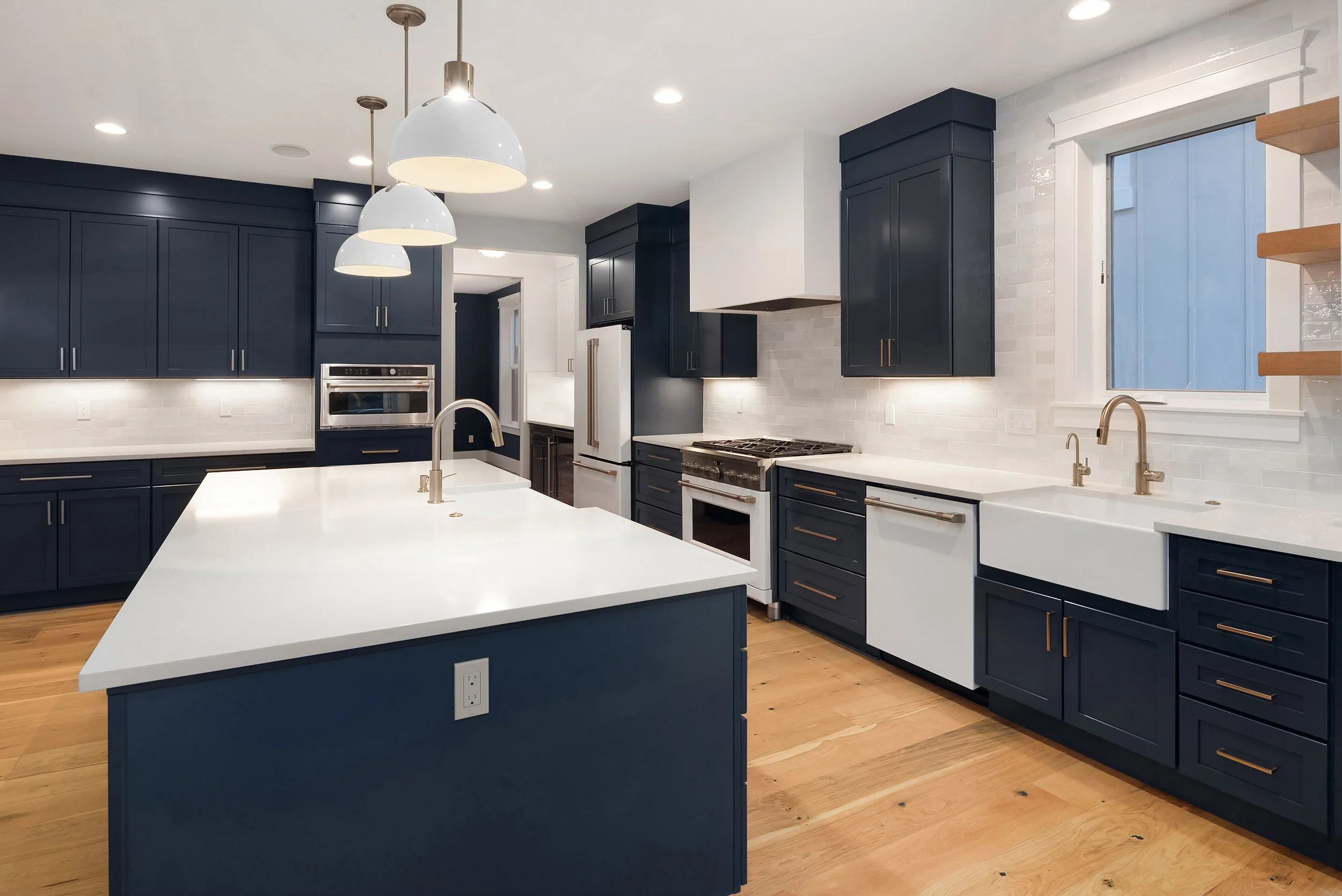 Modern kitchen with navy blue cabinets, white countertops, and a large island in the center. Features include stainless steel appliances, a white farmhouse sink, wooden flooring, and pendant lights hanging above the island.