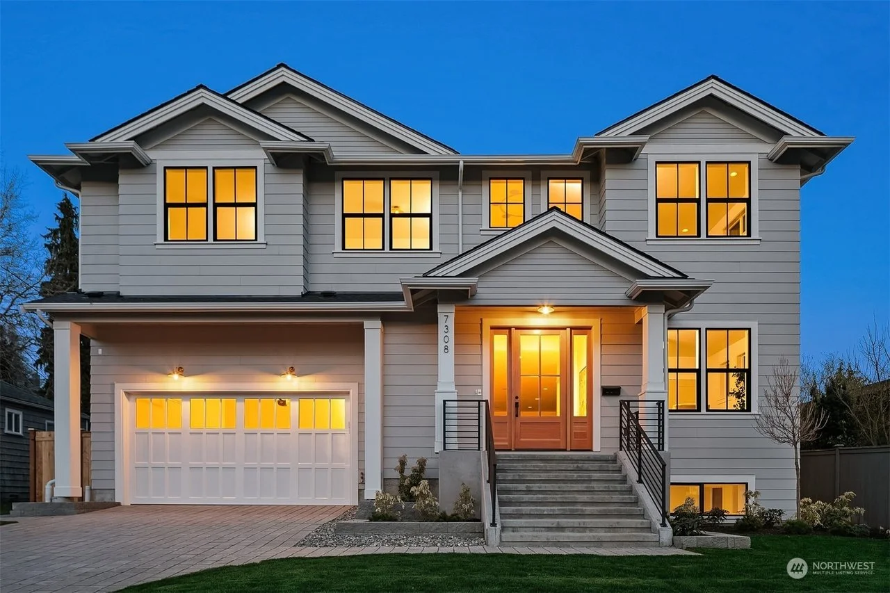 A modern two-story house with gray siding, illuminated windows, and a brightly lit front door at dusk. The house has a white garage door, a small front porch with stairs, and a well-maintained lawn.