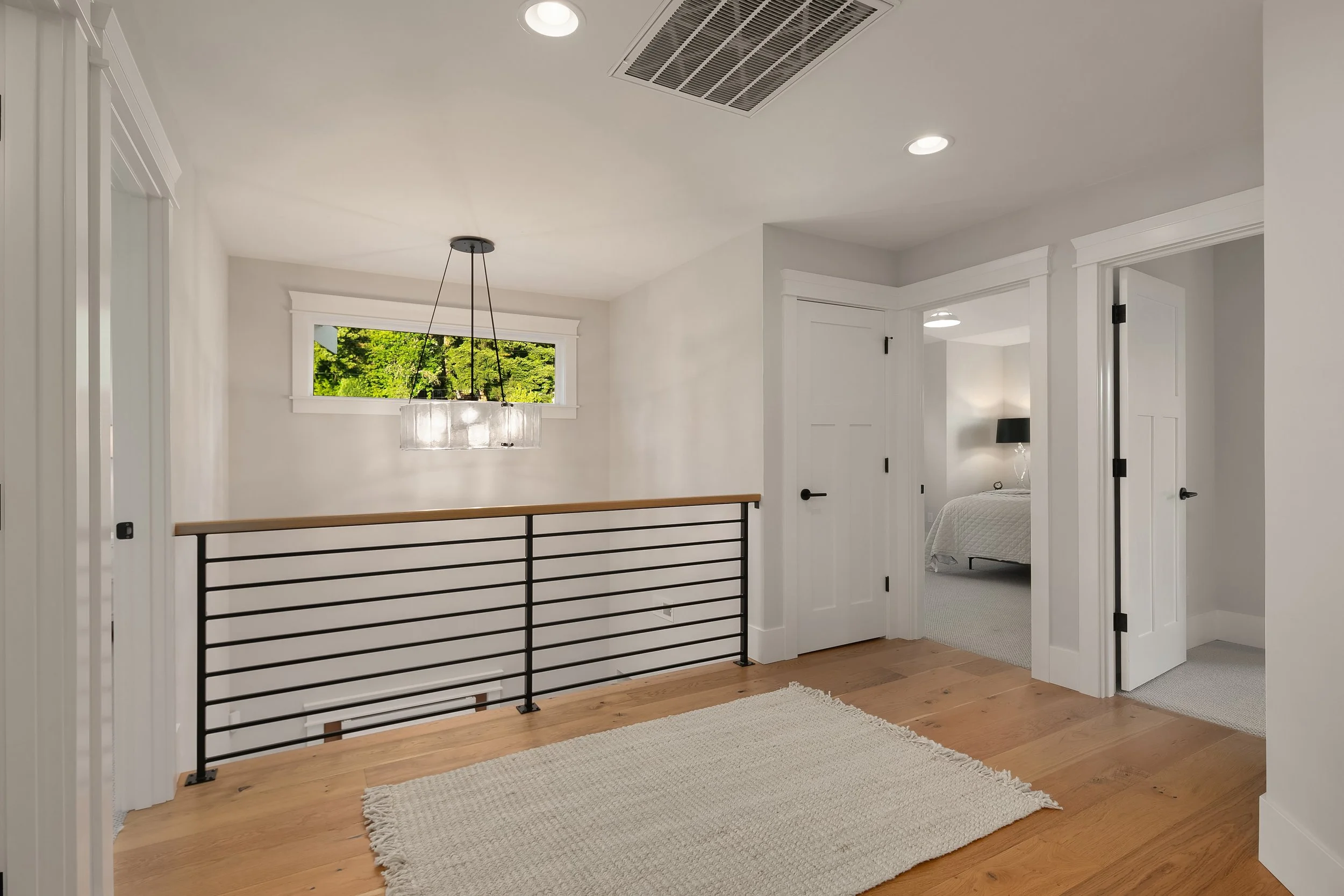 A bright upper landing in a modern home with white walls, hardwood floors, a small high window showing greenery outside, a black metal and wood railing, a modern pendant light, and two open doors leading to two bedrooms; one bedroom with visible bed 