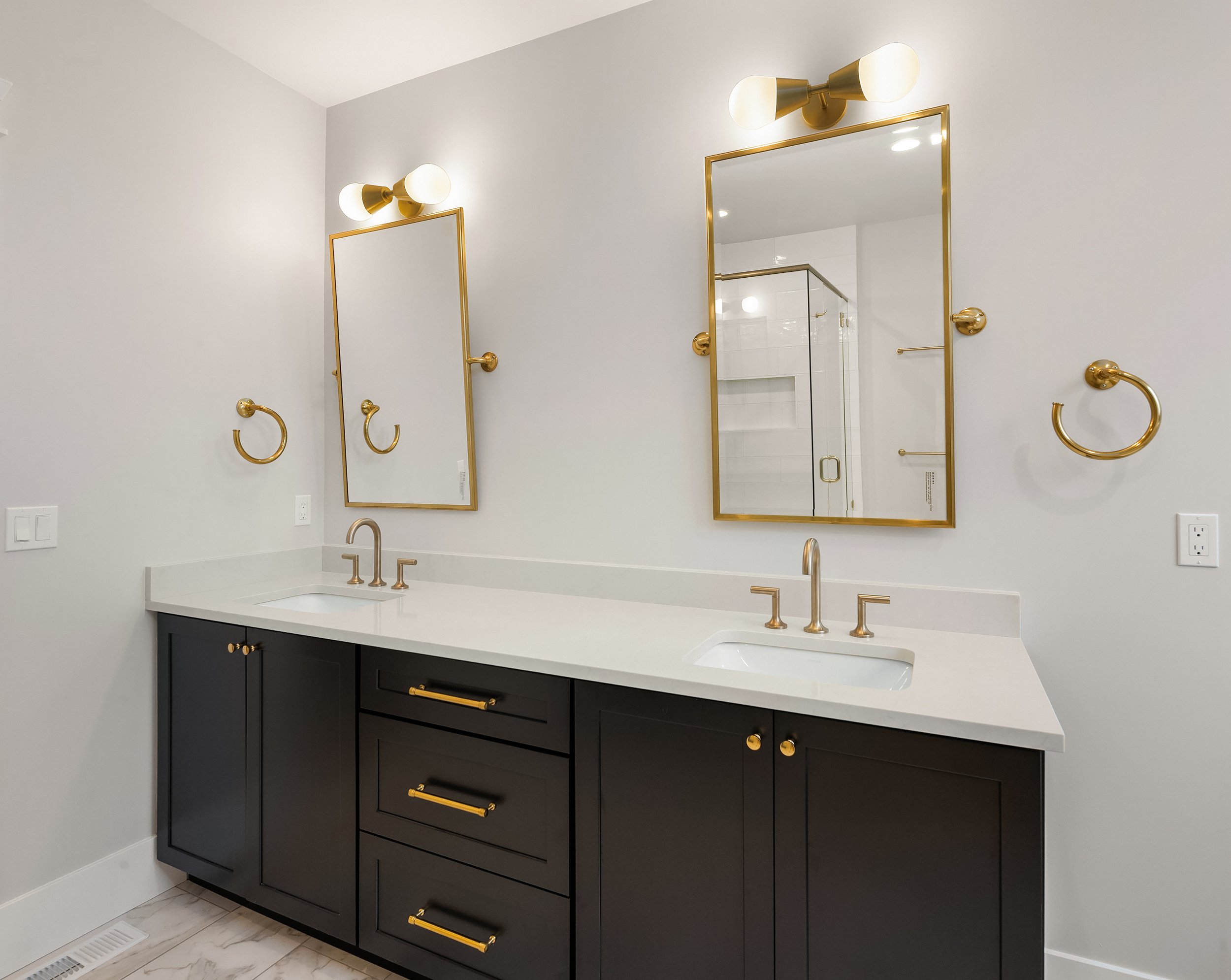 Bathroom vanity with two mirrors, black cabinets, white countertop, and gold fixtures, including faucets, handles, and towel rings, in a modern style.