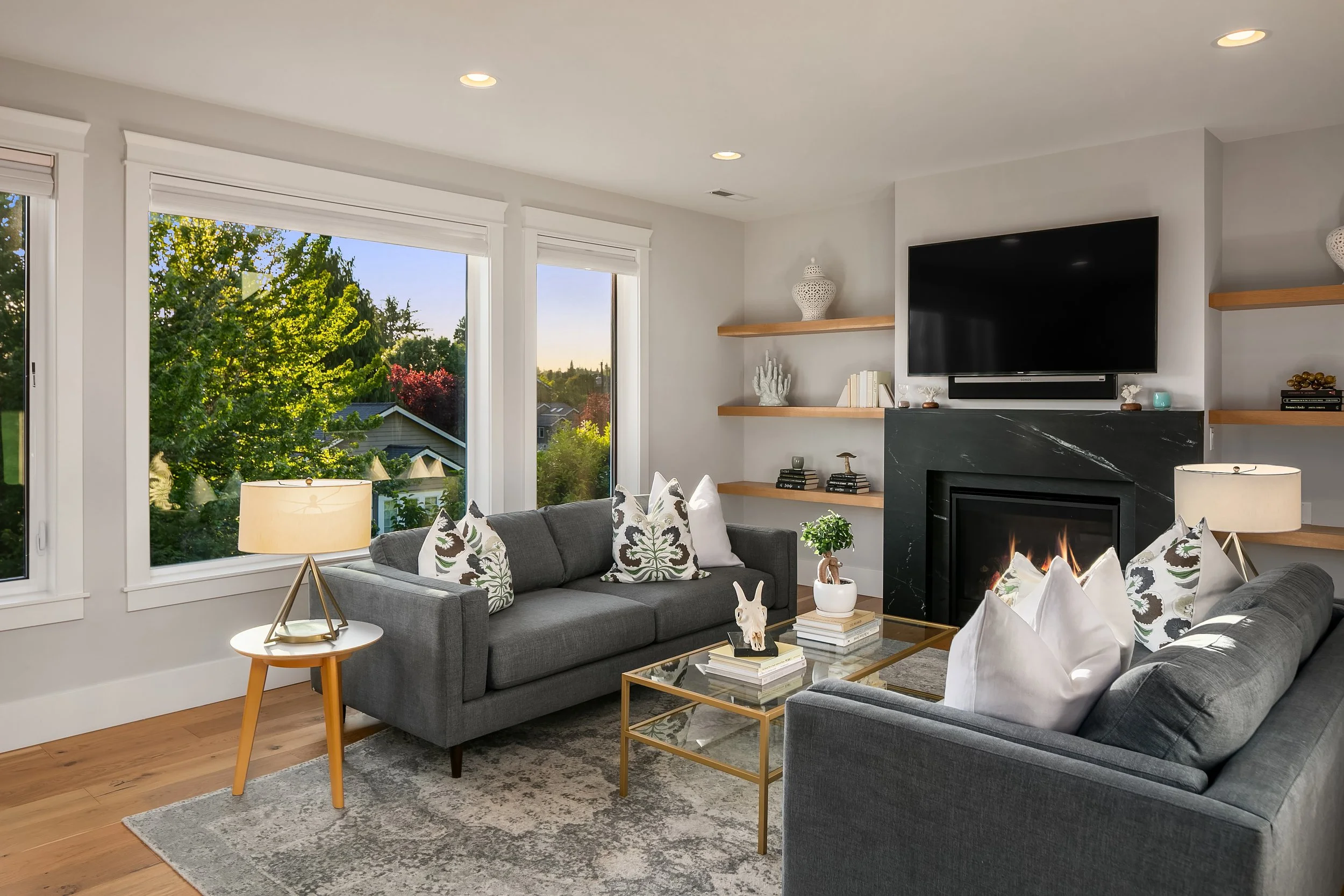 Living room with two dark gray sofas, white cushions, wooden shelves, a black fireplace, a TV, large windows showing green trees outside, and a glass coffee table with decorative items.