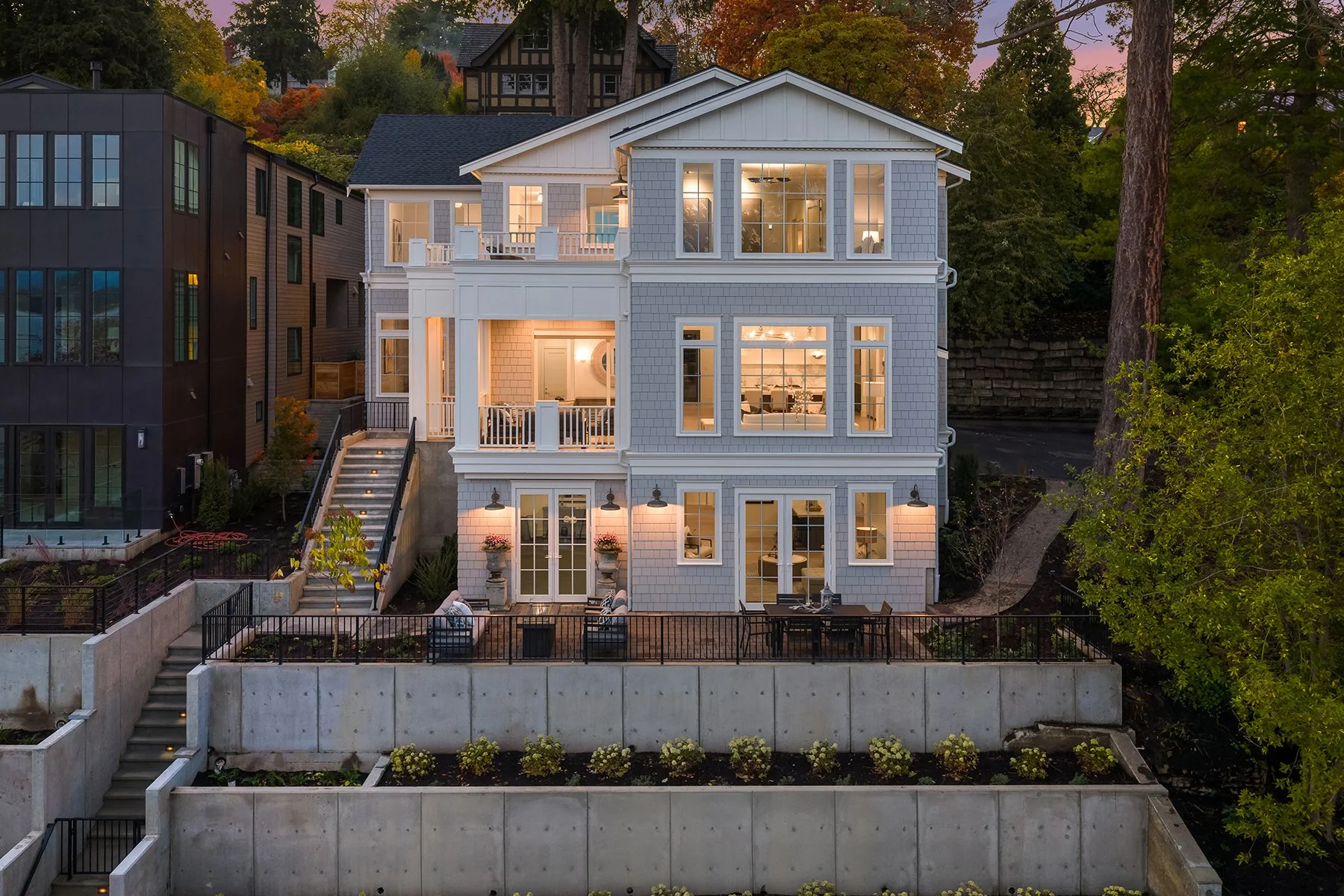 Nighttime view of a multi-story house with large windows, illuminated from within, situated on a terraced landscape with a staircase and outdoor seating areas, surrounded by trees with fall foliage.