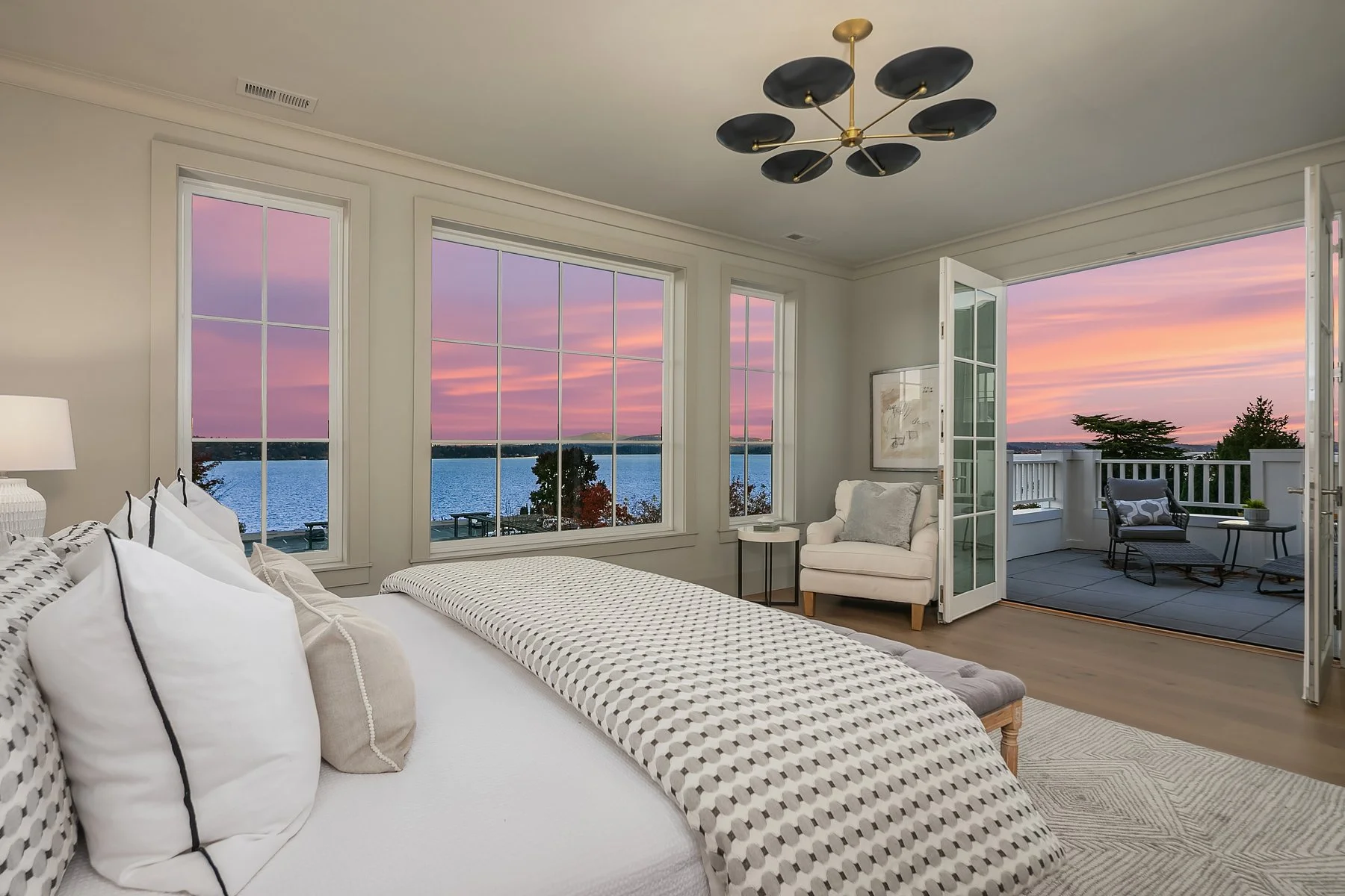 Bedroom with large windows and a view of a lake and sunset, featuring a bed, white armchair, side table, ceiling light fixture, and outdoor balcony.