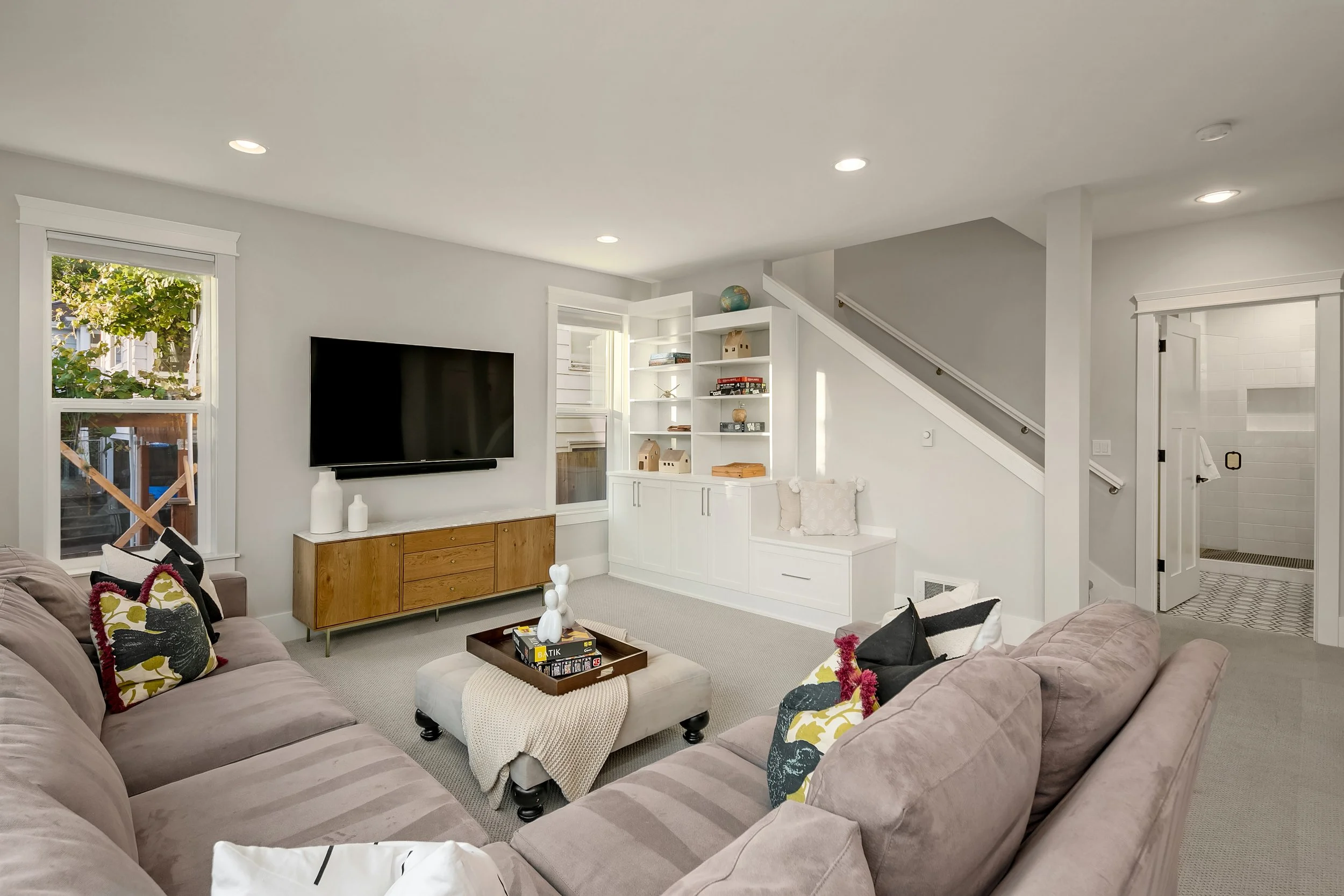 Living room with beige sofa, black and white pillows, a wooden TV stand, a wall-mounted television, built-in white bookshelves, staircase with white risers, light-colored walls, and a doorway leading to a bathroom with white tiles.