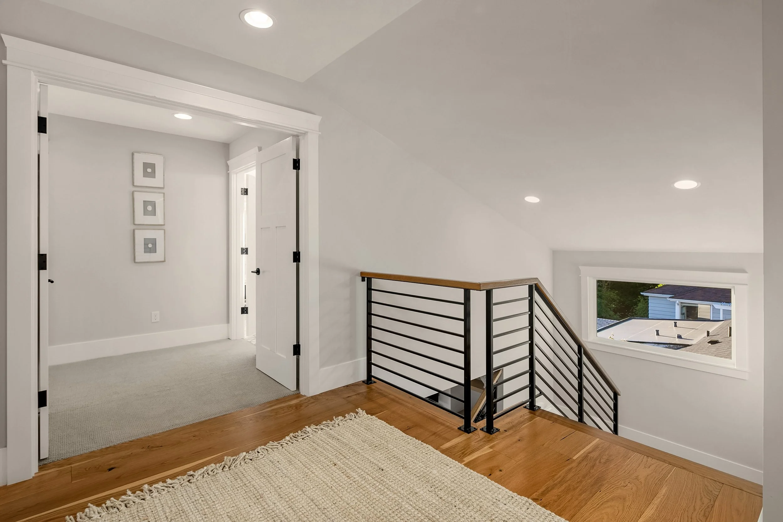 Interior of a contemporary house's upper floor landing with white walls, wooden flooring, a black metal railing with wooden top, a window looking out to neighboring houses, and a doorway leading to a room with carpeted flooring and three framed pictu