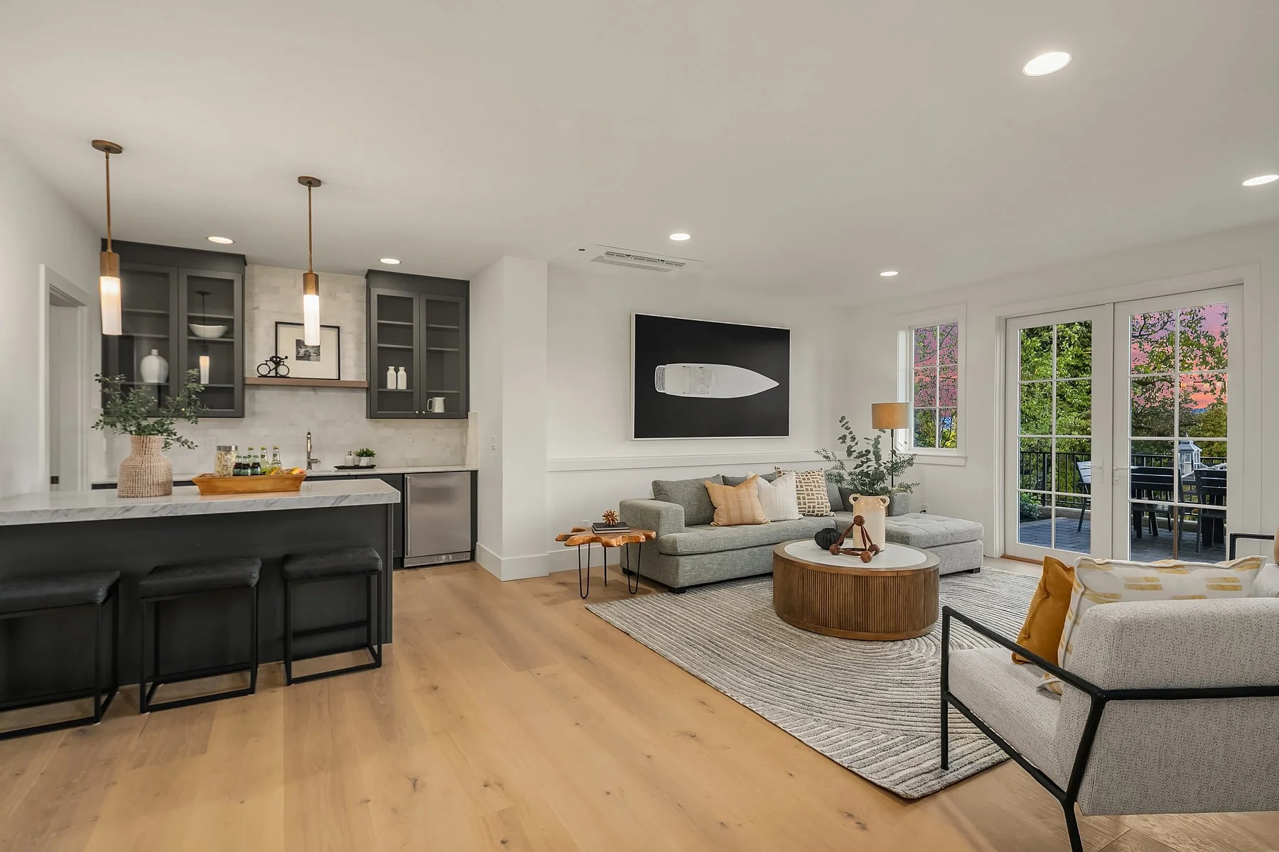 Living room with gray sofa, armchair, wood coffee table, and sliding glass doors showing outdoor patio with trees and pink sunset sky.