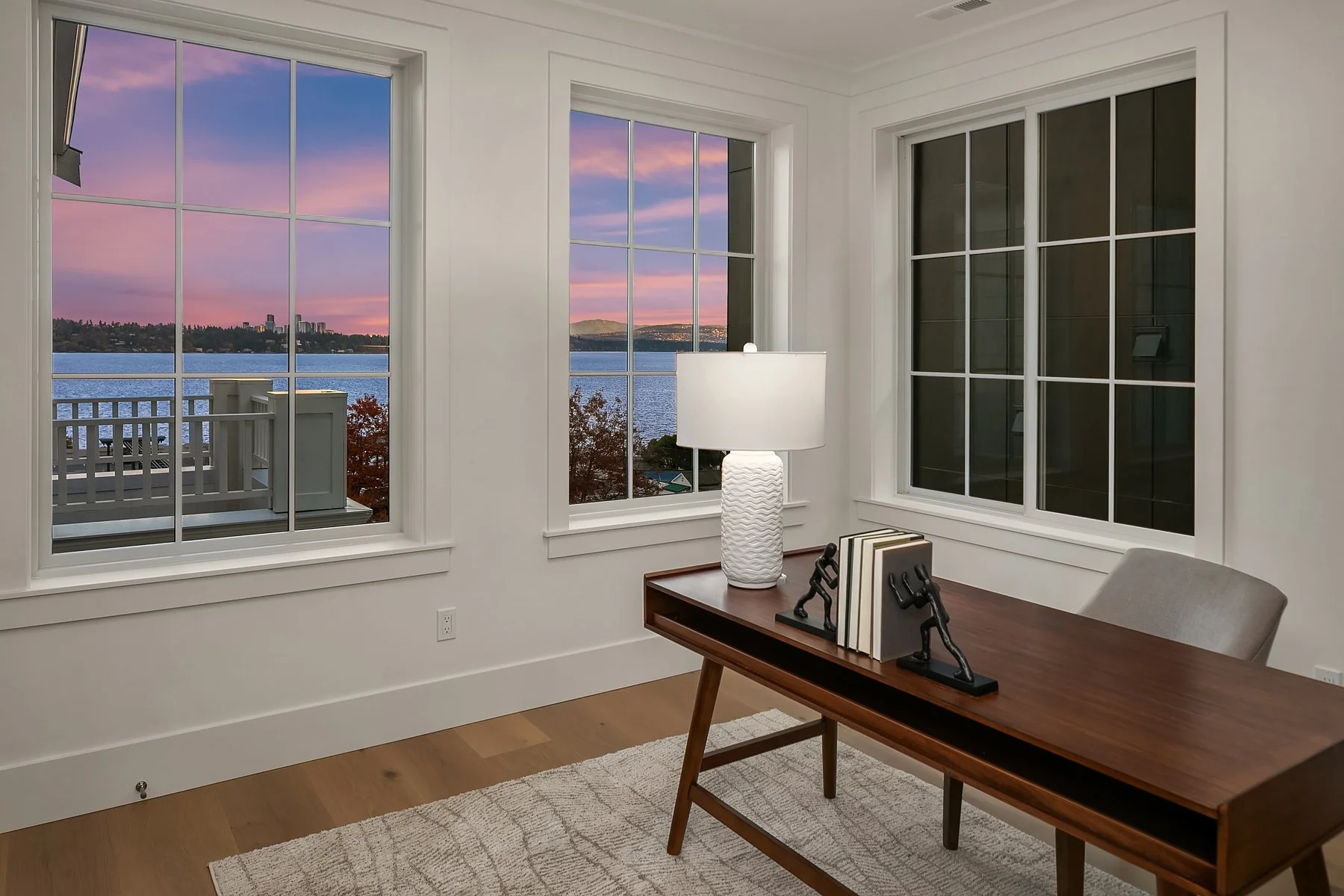 Interior of a room with three large windows showing a scenic view of a body of water, trees, and distant buildings at sunset. The room has white walls, a wooden table with decorative figures and books, a table lamp, and a light-colored carpet.