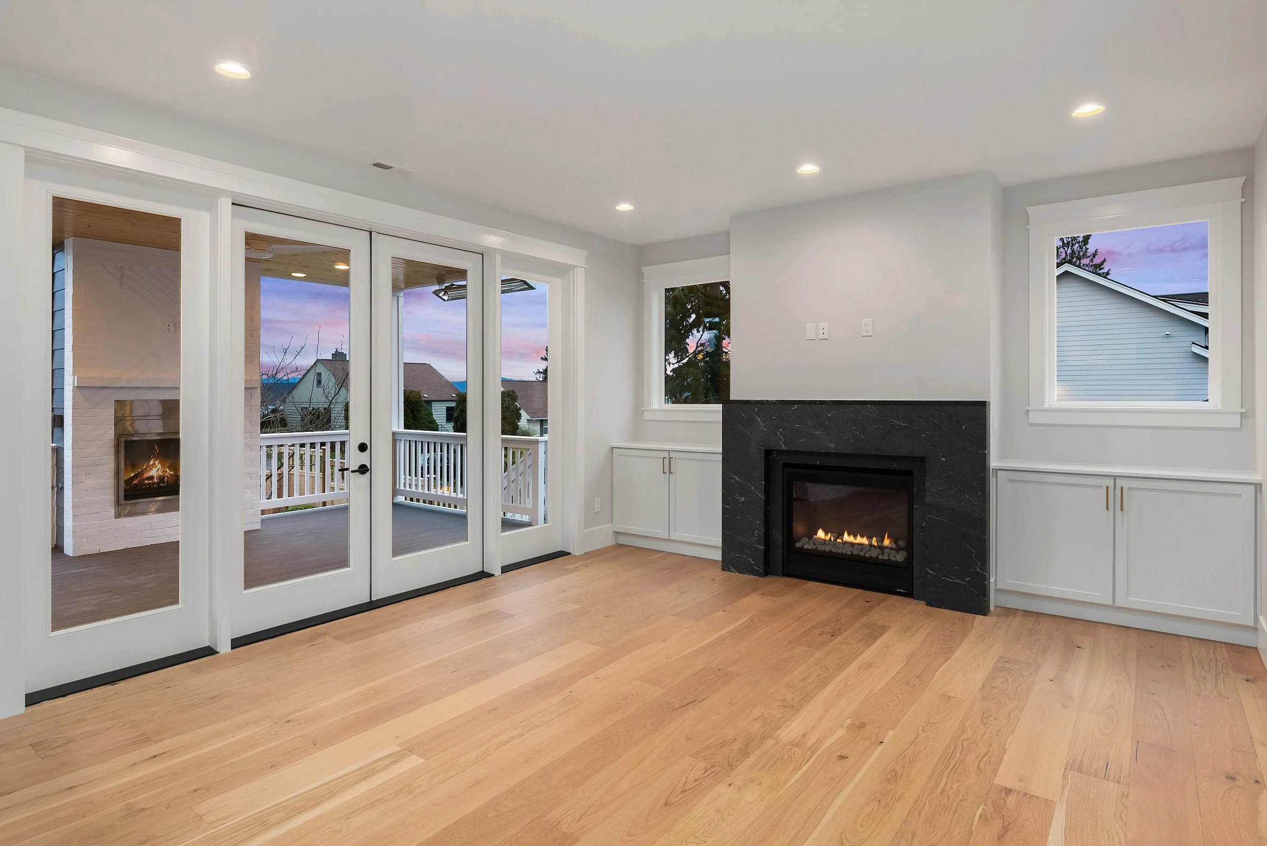 Empty living room with hardwood floors, white walls, large windows, a black fireplace, and a sliding glass door leading to a balcony with a fireplace outside.
