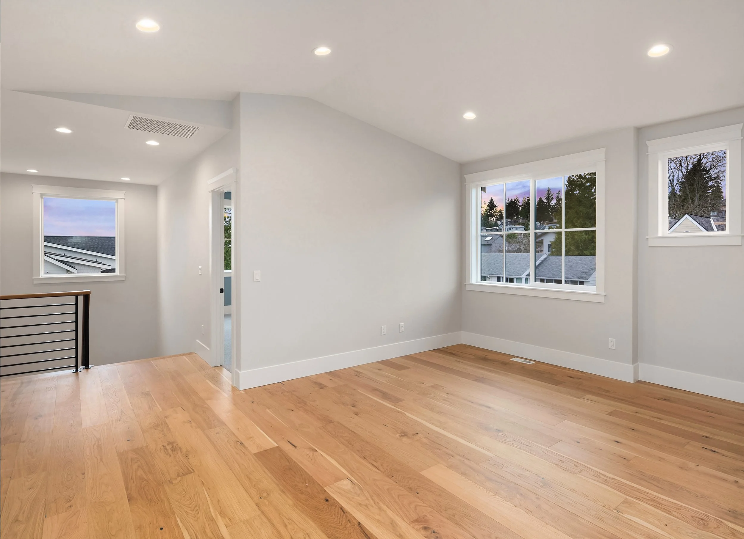 Empty room with white walls, hardwood floors, and multiple windows showing a neighborhood view at sunset.