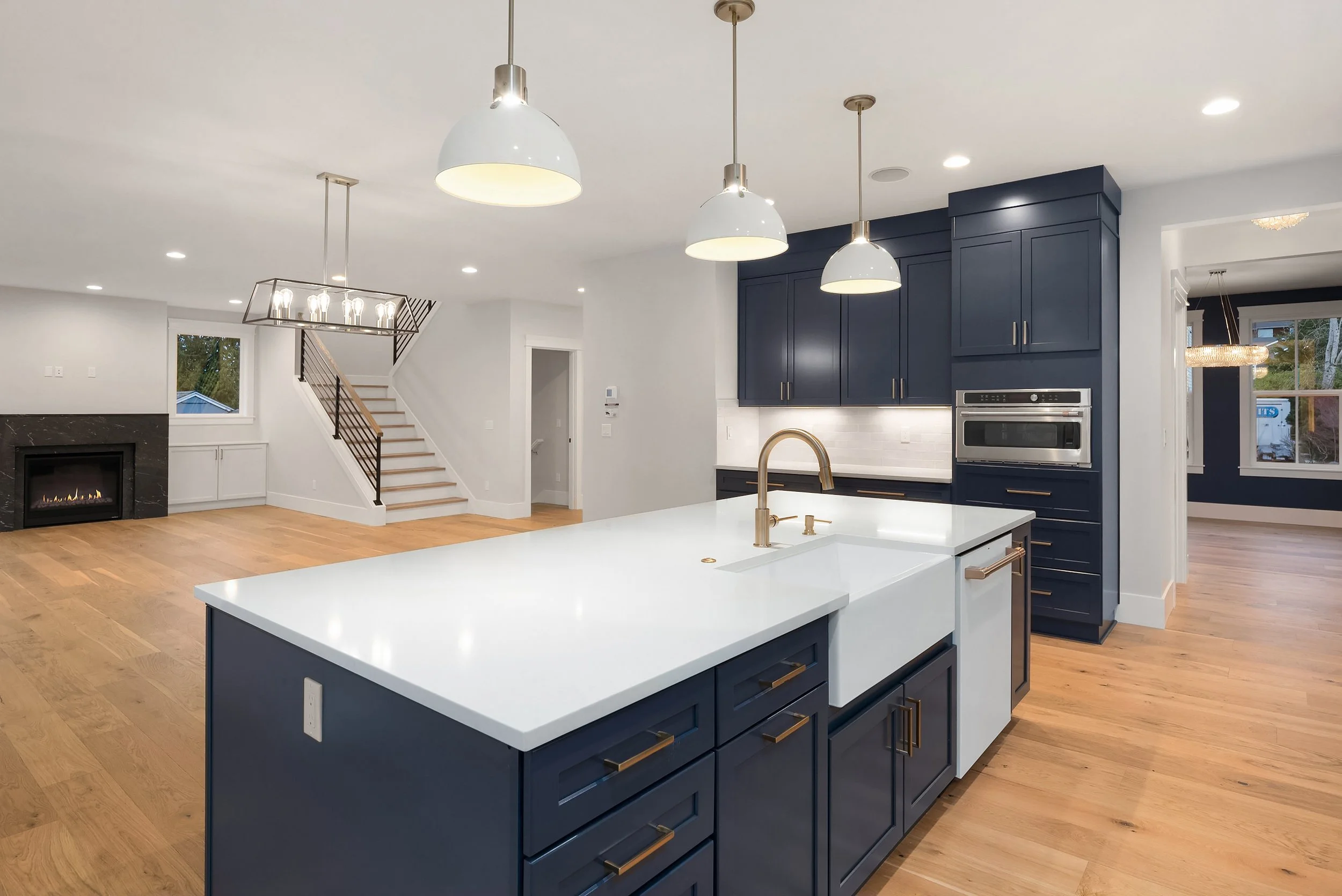Modern kitchen with navy blue cabinets, white countertop, and a farmhouse sink. Open living area with a fireplace, wooden floors, and stairs leading upstairs. Pendant lights hang over the kitchen island.