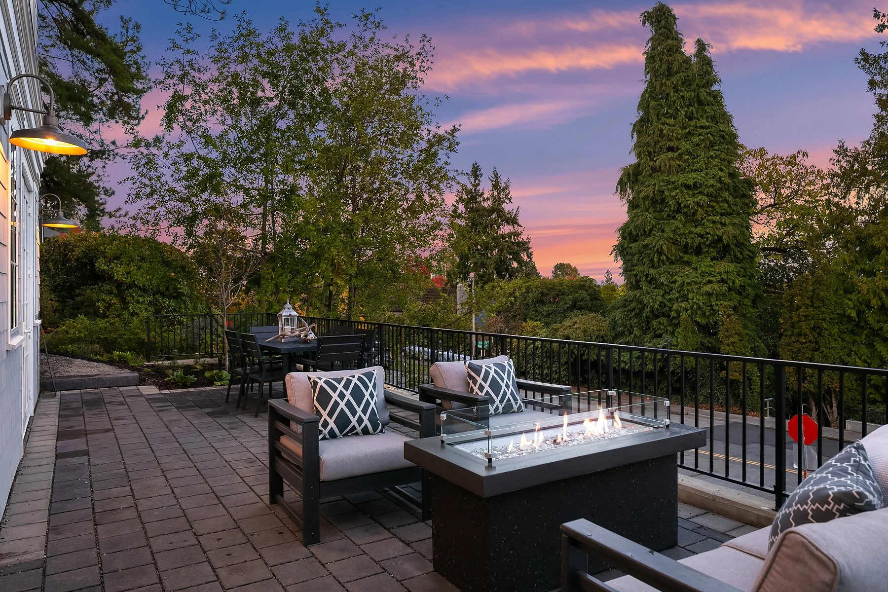 Outdoor patio with seating area, modern fire pit, dining table, trees, and evening sky with pink and purple clouds.