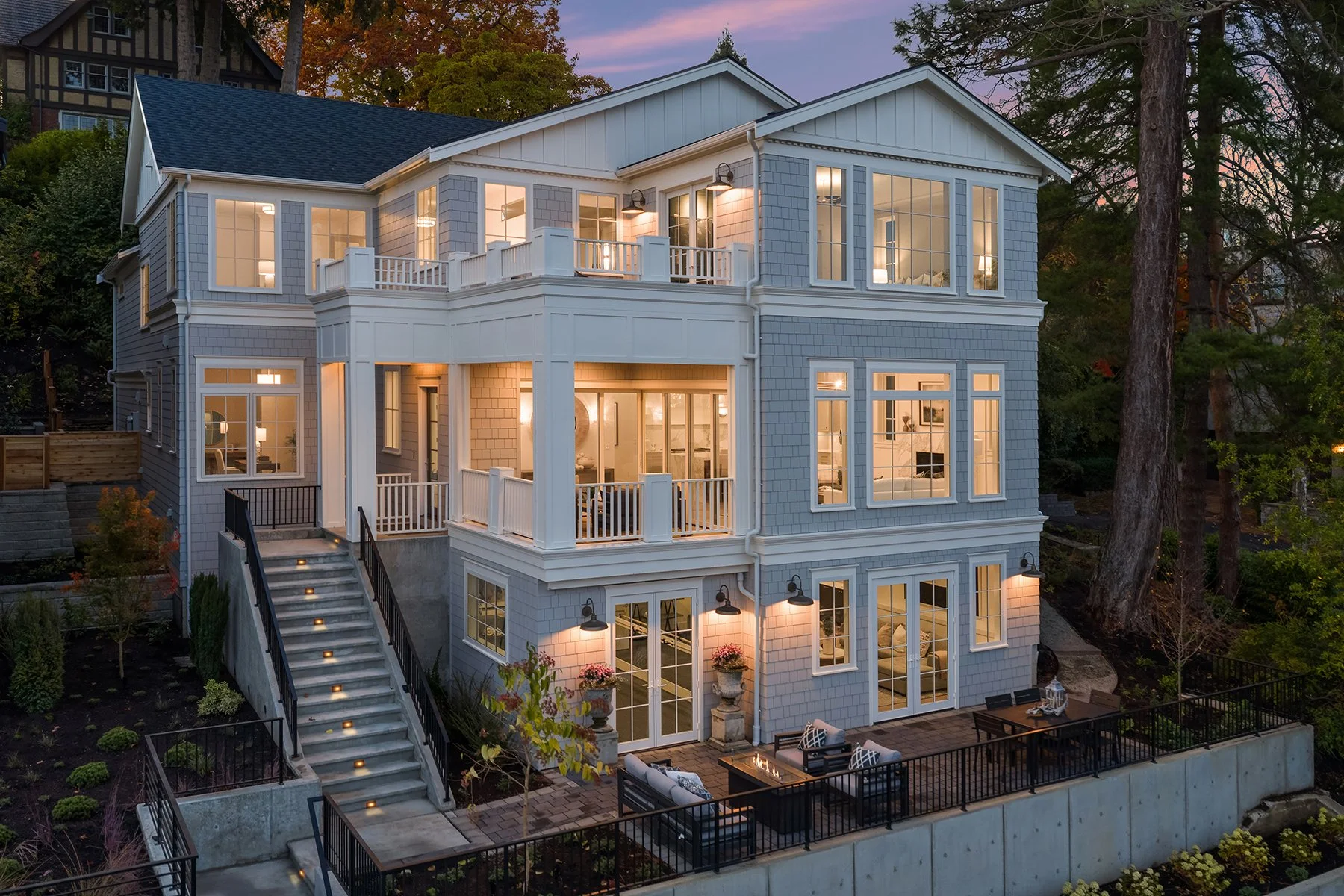 A modern multi-story house with large windows, a spacious deck, and outdoor seating, illuminated at dusk.