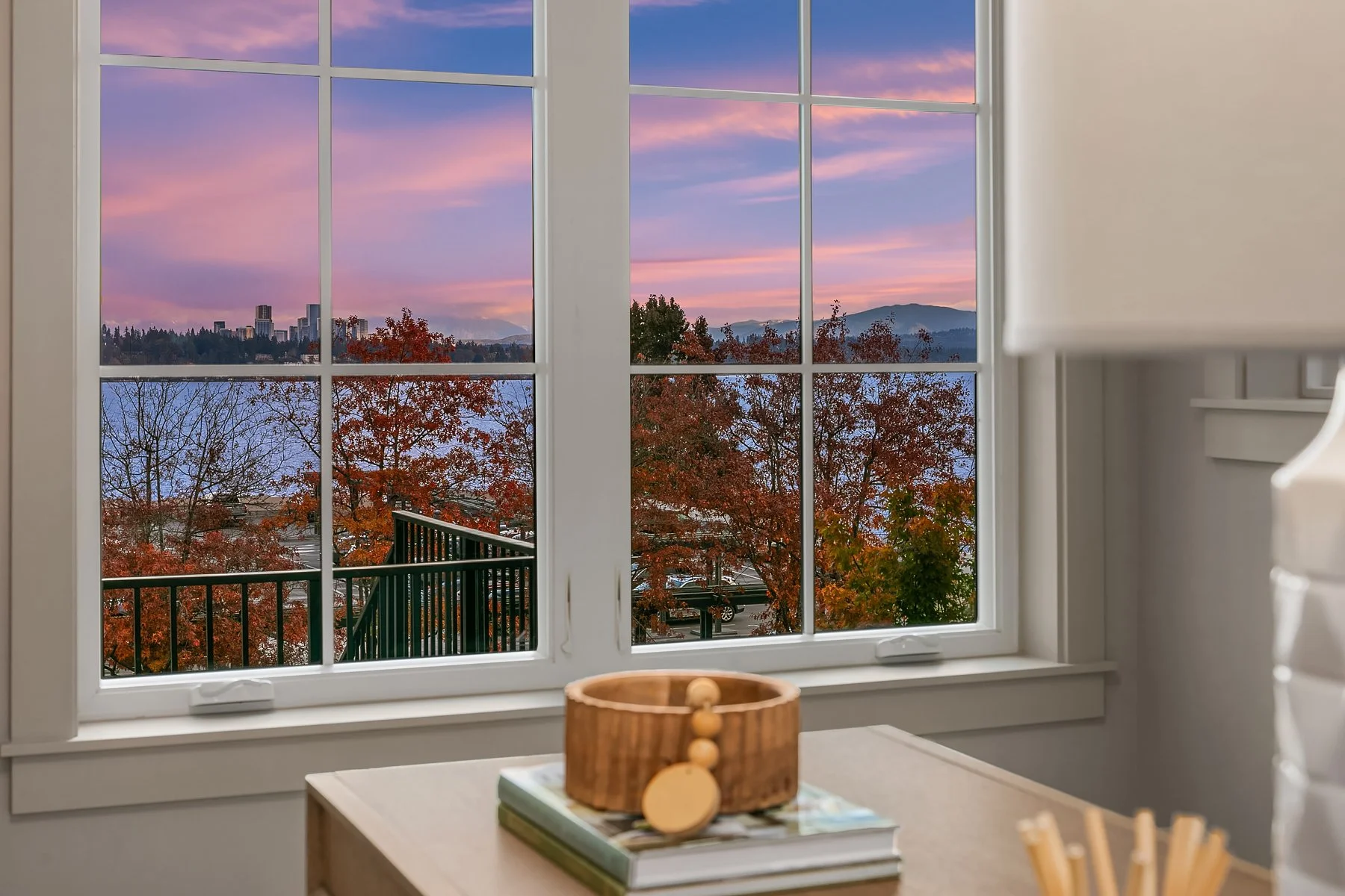View of an outdoor scene through a white-framed window showing trees with red and orange leaves, a body of water, a city skyline, and mountains in the distance at sunset.