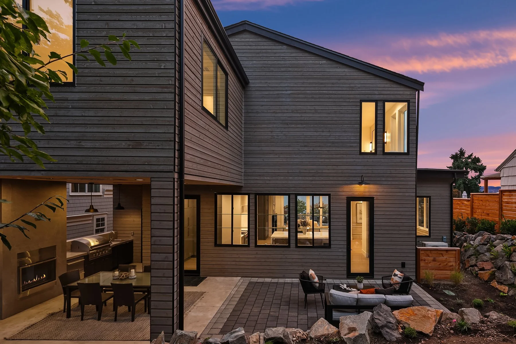A two-story modern house with wooden siding during sunset, featuring large windows, a cozy outdoor seating area with a sofa and chairs, and a built-in fireplace on the left side.